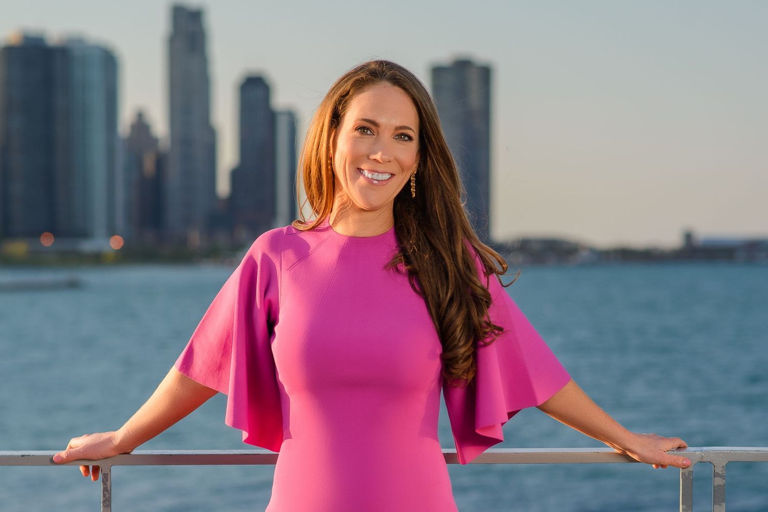 Someone in a bright pink dress stands on a boat deck with the Chicago skyline visible across the water at sunset.