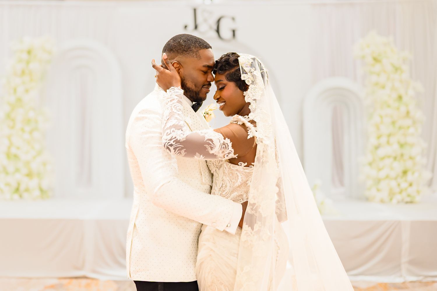 Bride and groom share first dance in elegant white wedding attire before floral arch backdrop.