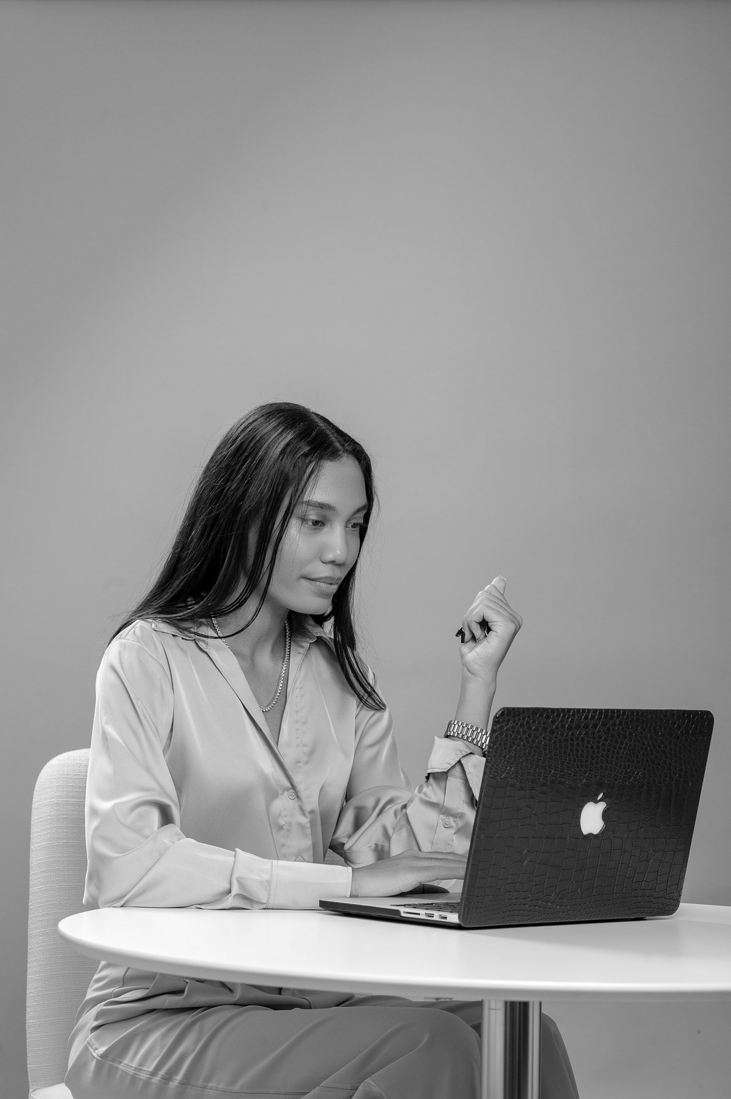 Woman in silk blouse working on Apple MacBook laptop at white round table, black and white professional photo.