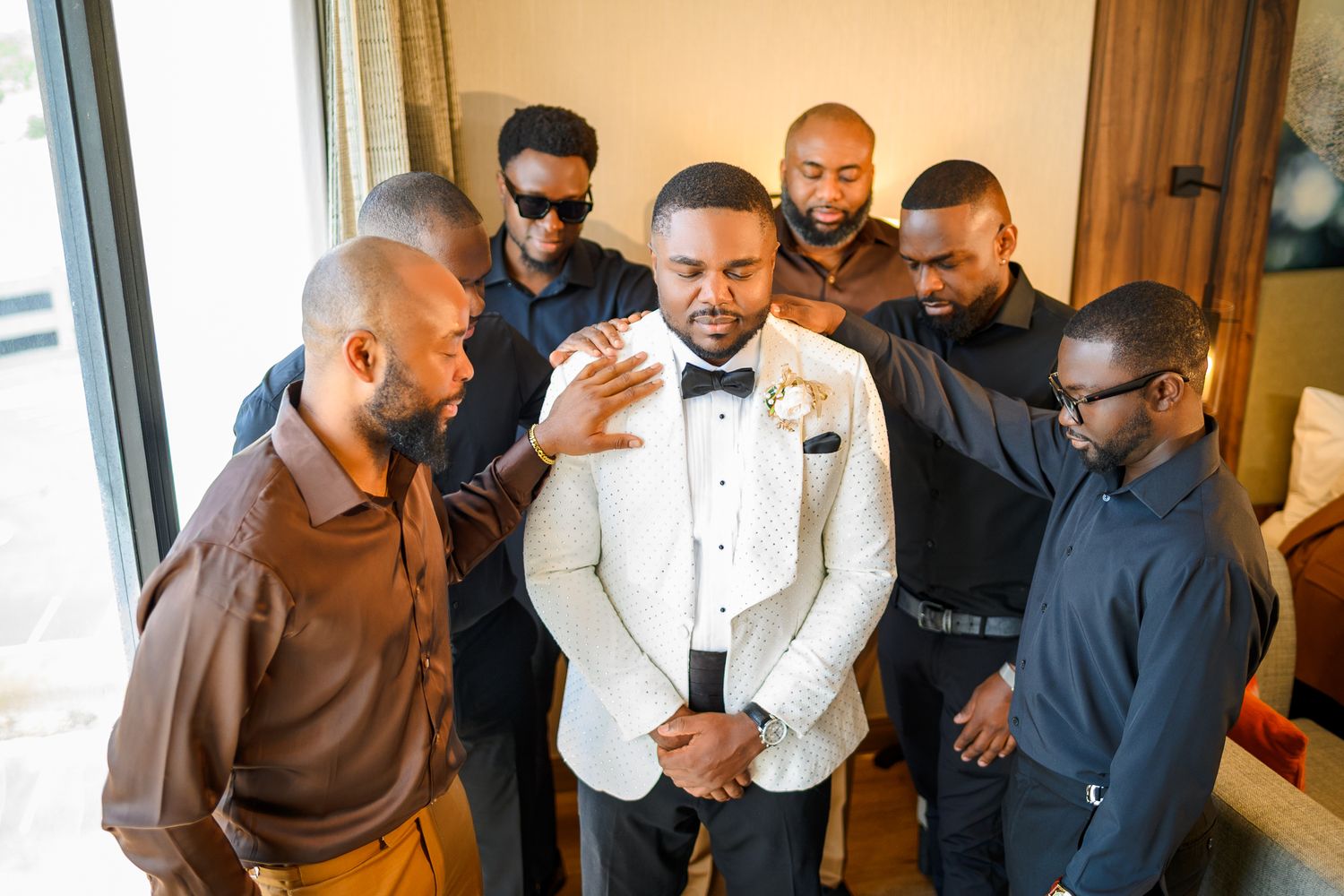 Groom in white tuxedo jacket surrounded by groomsmen adjusting his bow tie before wedding ceremony.