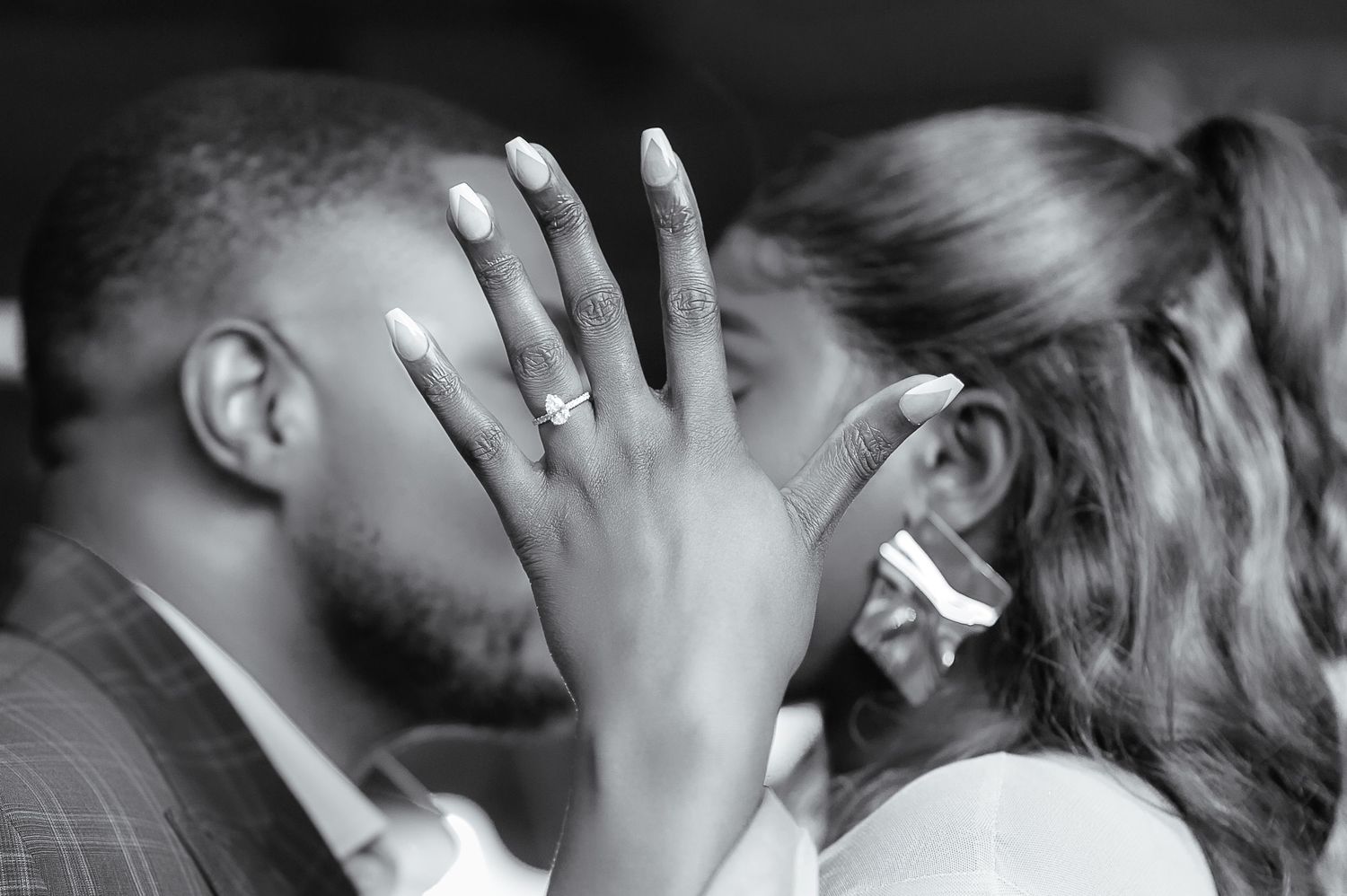 Black and white photo of a couple kissing while the woman displays her engagement ring to the camera.