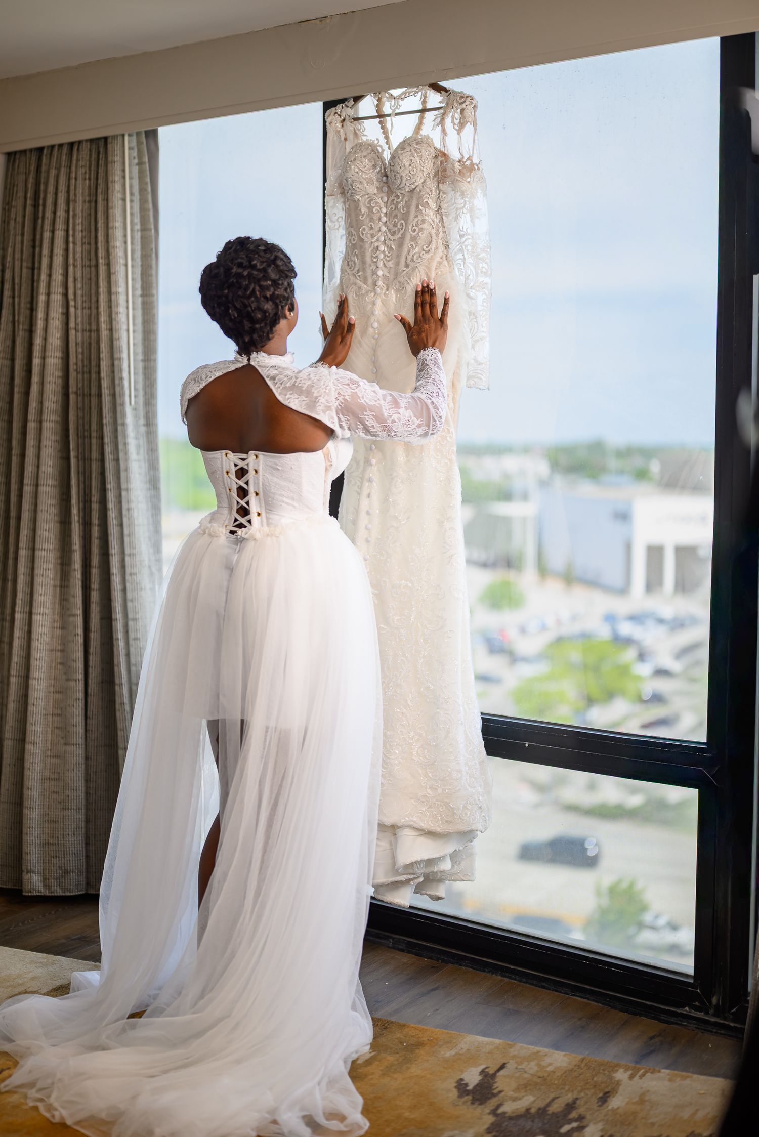 Bride in white tulle skirt and lace corset top reaches for wedding gown hanging by hotel window.