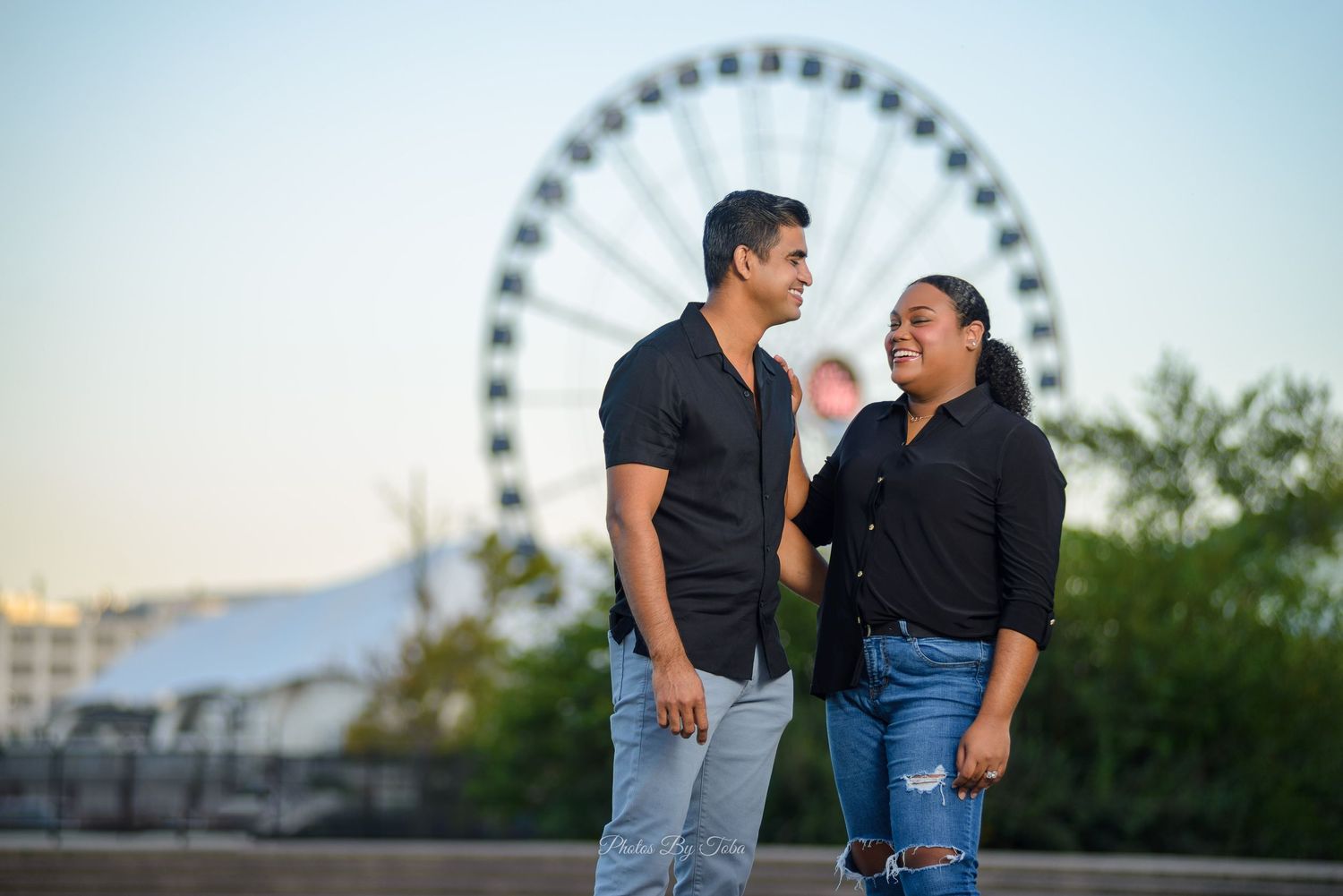 Couple smiling at each other near a ferris wheel at sunset, man in black shirt and jeans, woman in black top.