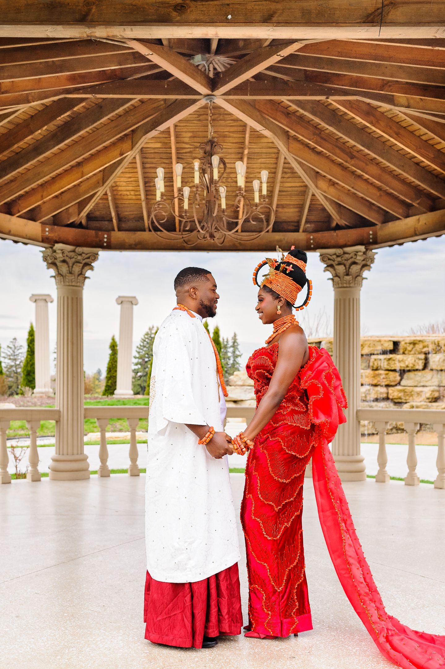 Couple in traditional Nigerian wedding attire posing under elegant gazebo, bride in red gown with orange headpiece.