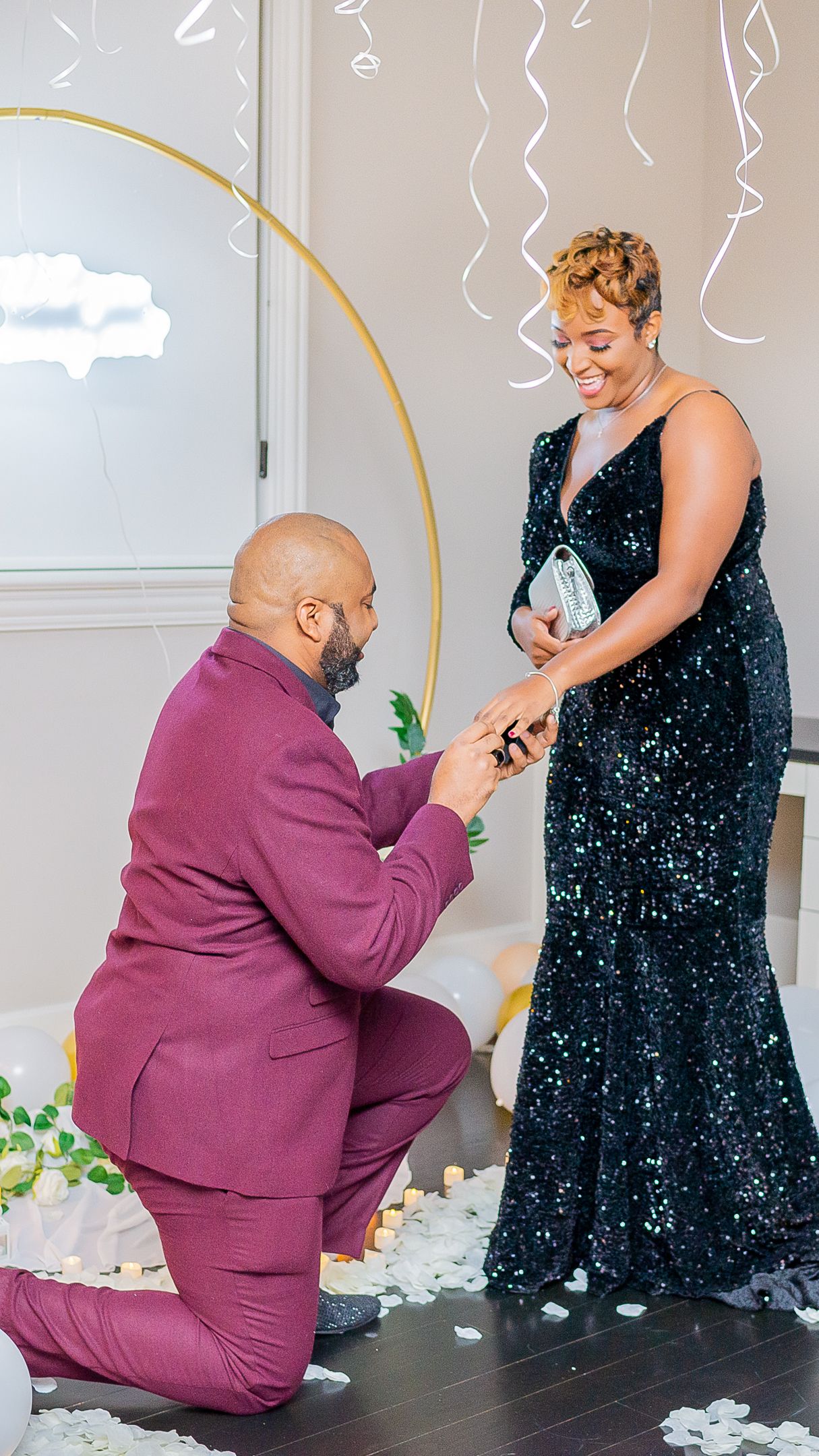 Man in burgundy suit proposes on one knee to woman in black sequin gown amid rose petals and streamers.
