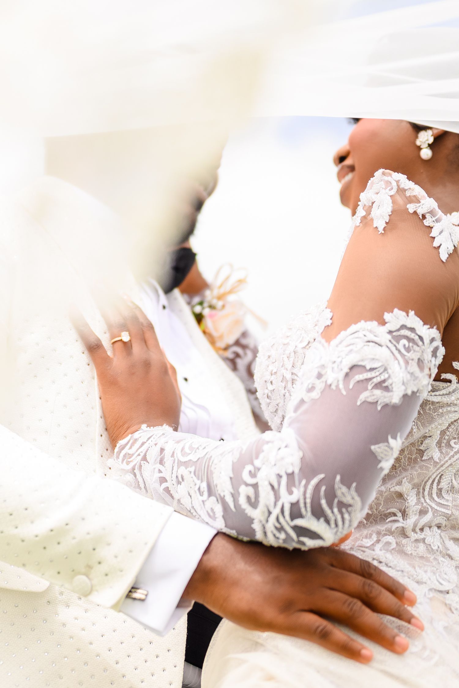 Bride in lace wedding gown embracing groom in white suit, showcasing elegant floral lace sleeves and diamond ring.