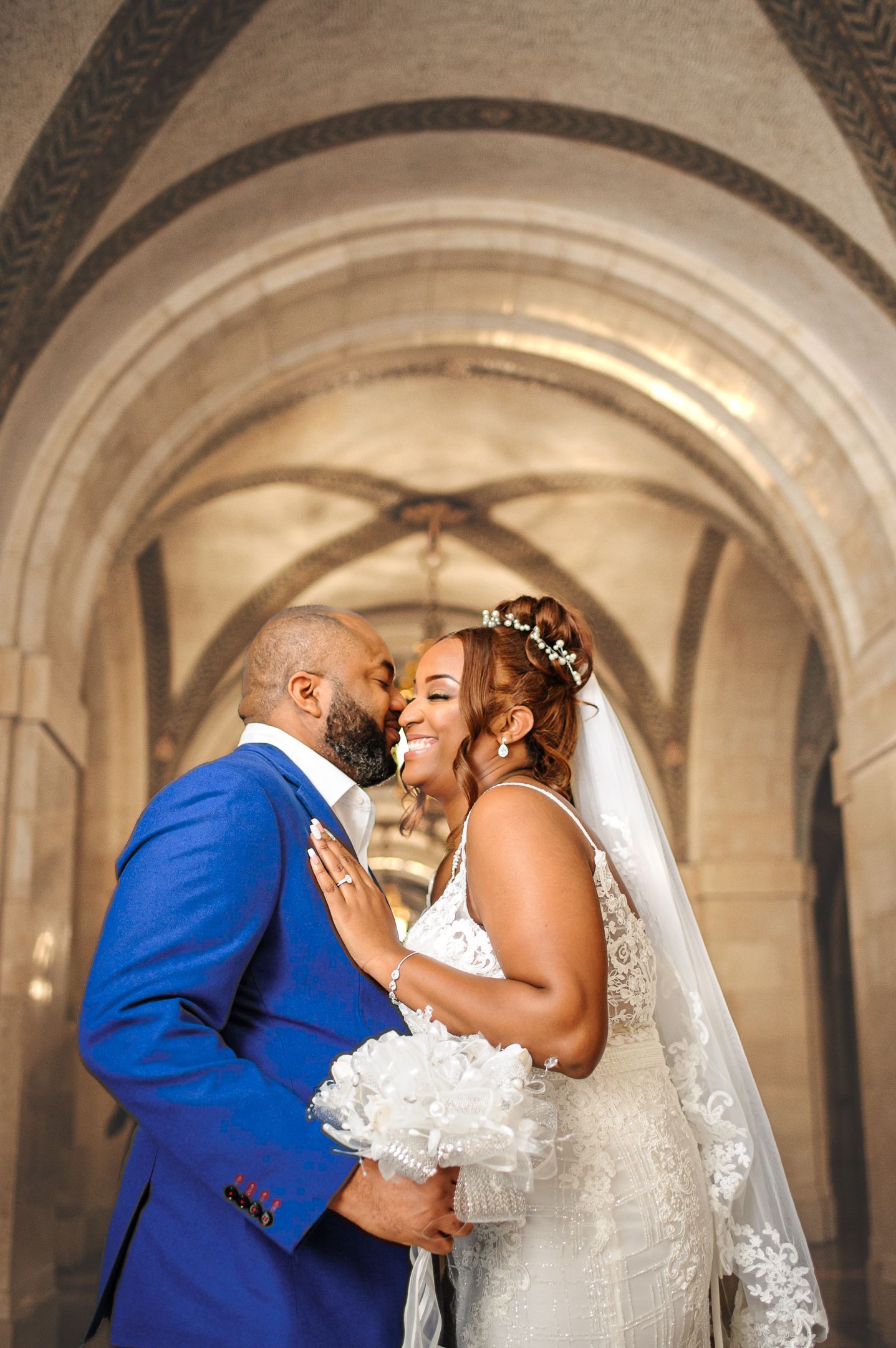 A bride and groom share a romantic moment under stone arched ceilings, she in a lace gown with veil, he in a blue suit.