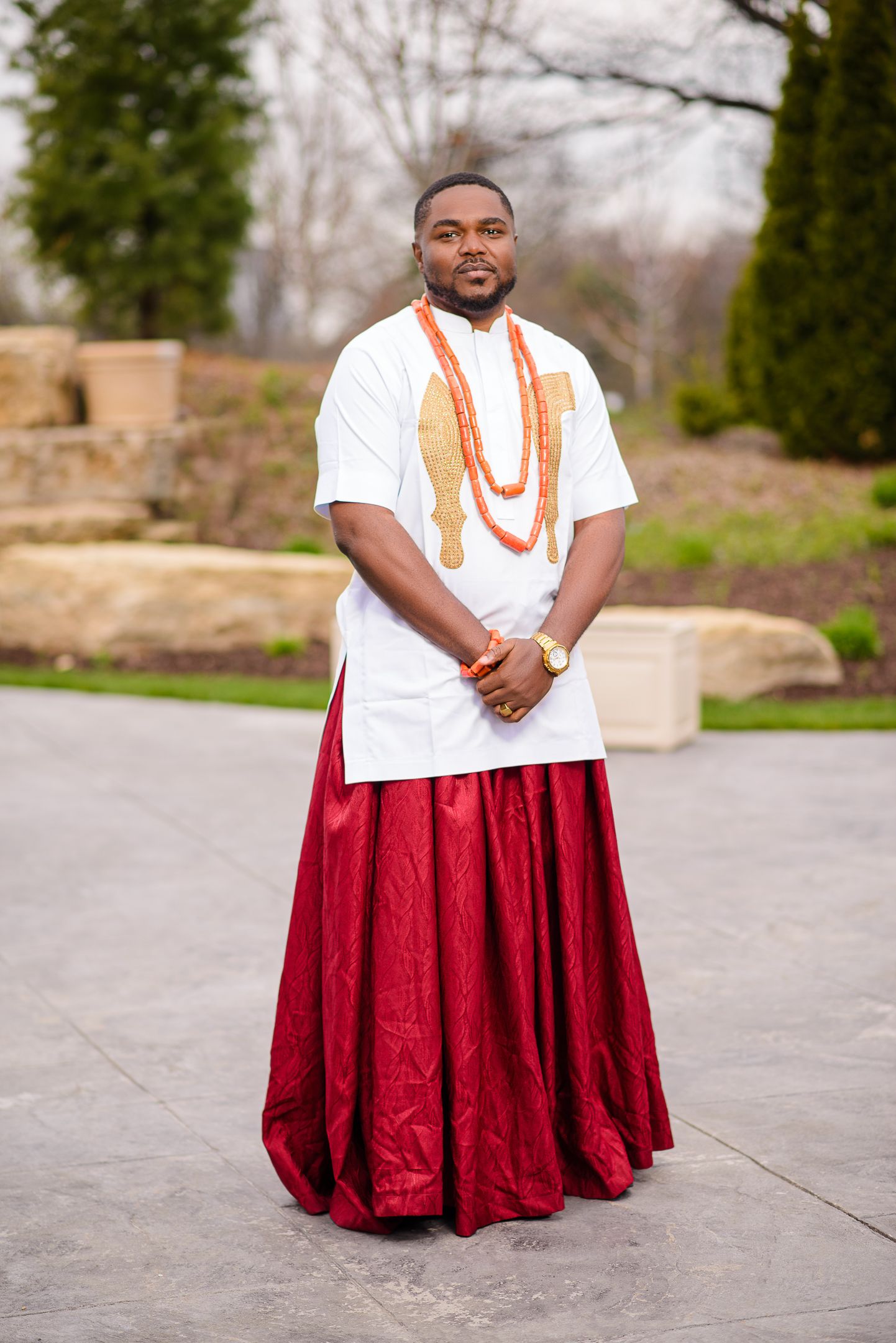 Nigerian groom in white embroidered top, red wrapper, and coral beads stands in garden.
