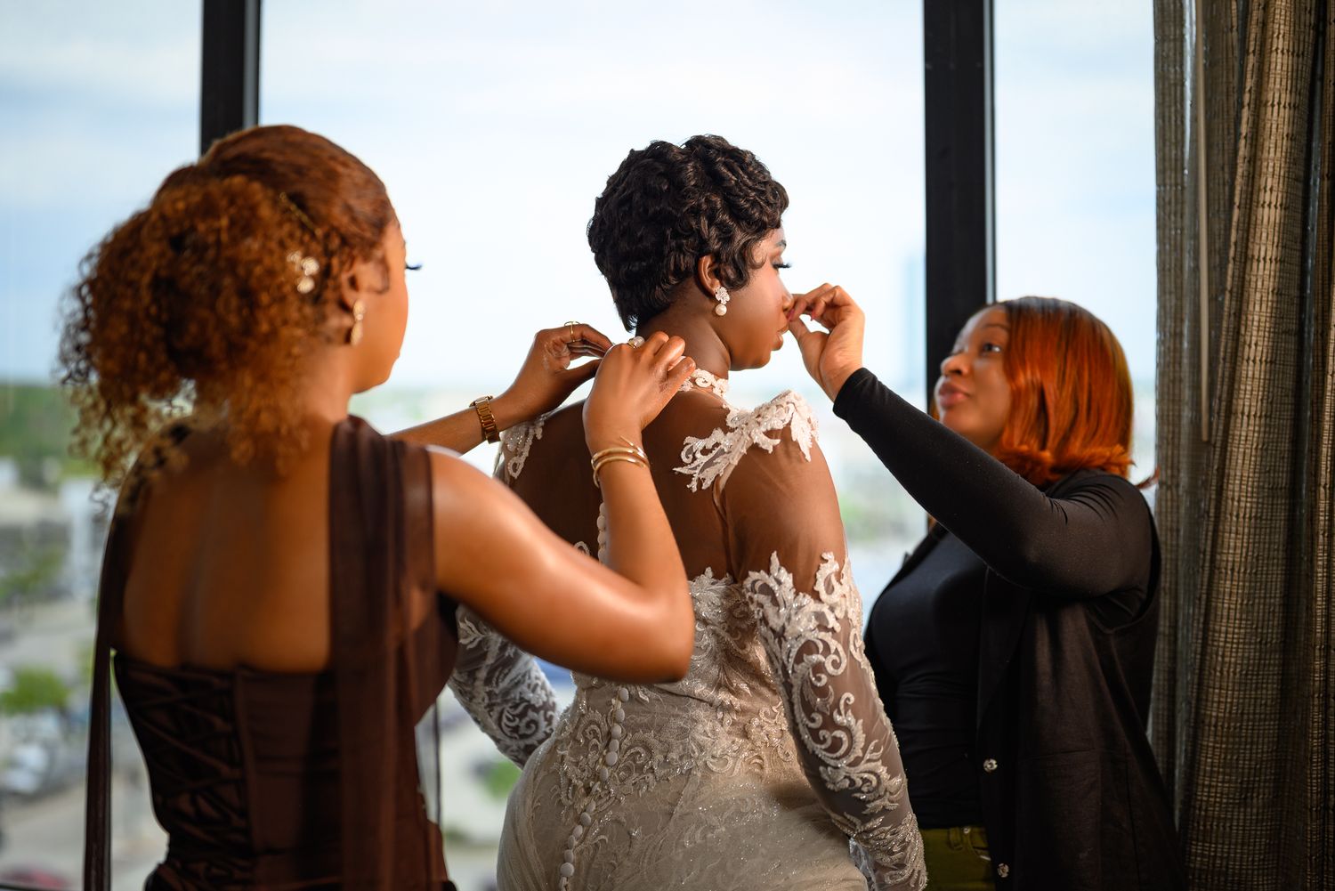 Bride in lace wedding gown being helped by two bridesmaids near a large window during bridal preparations.