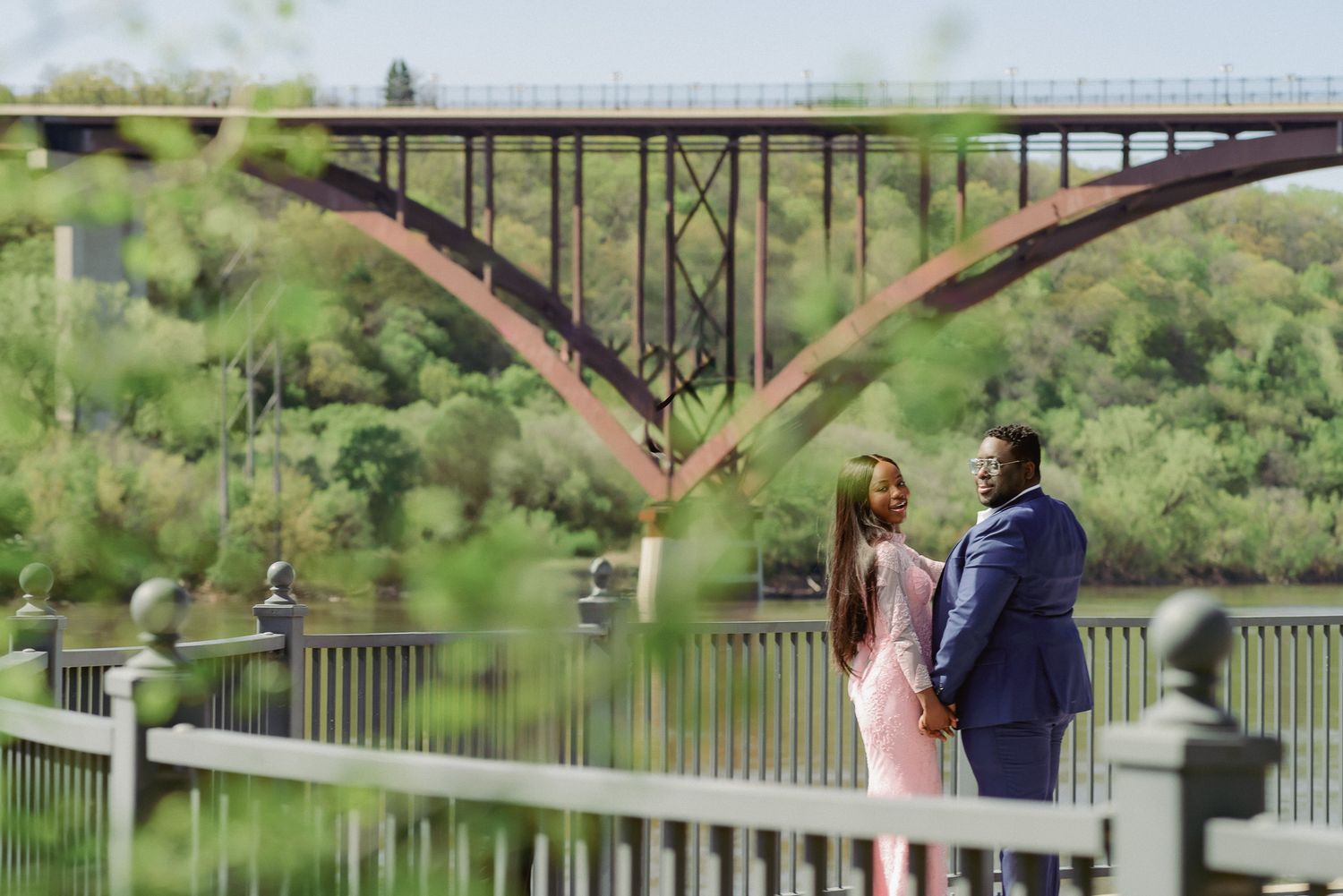 Couple in formal wear sharing a romantic moment on a bridge overlooking a river with a red arch bridge backdrop.
