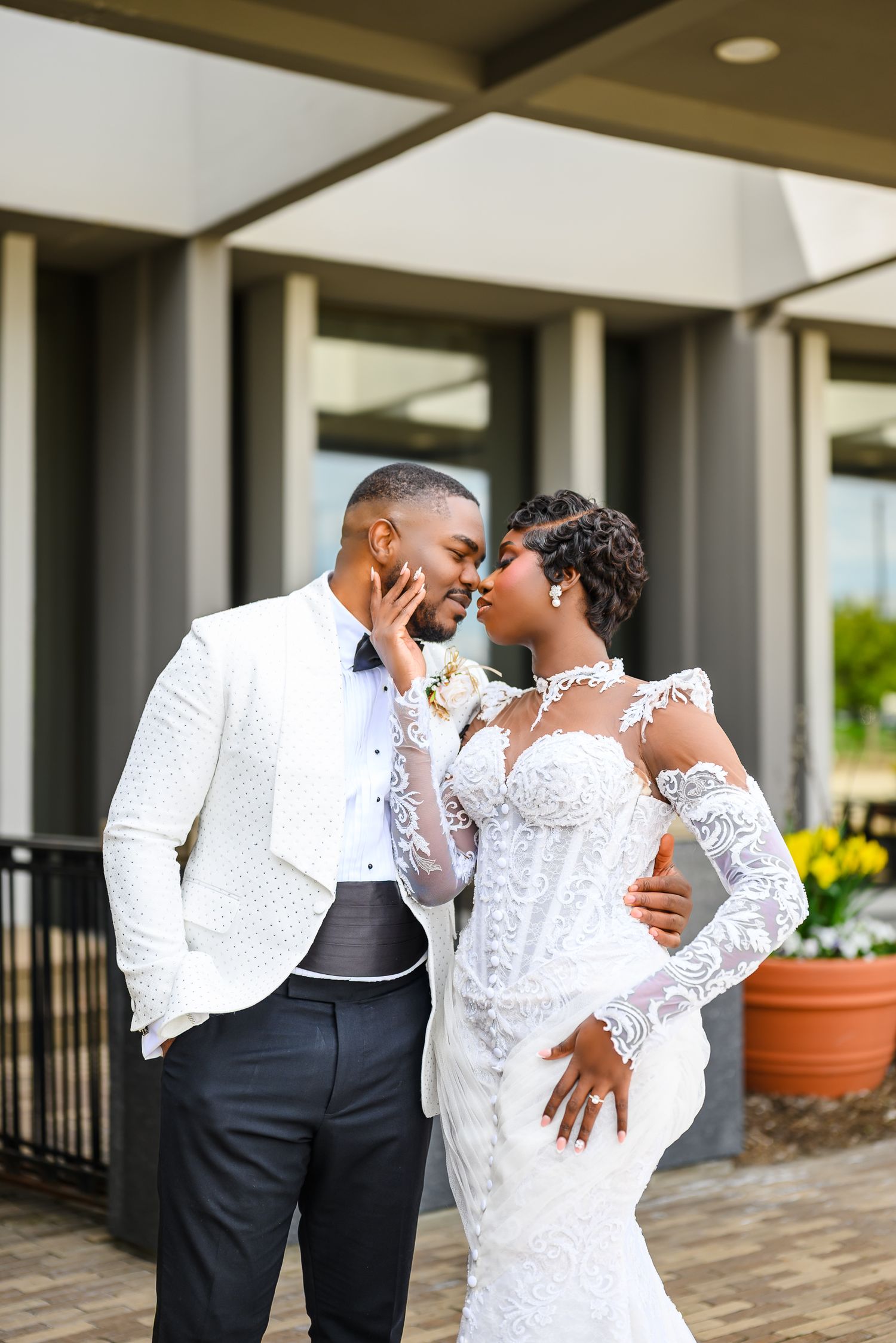A Black bride and groom share a romantic moment in wedding attire, she in lace gown, he in white tuxedo jacket.