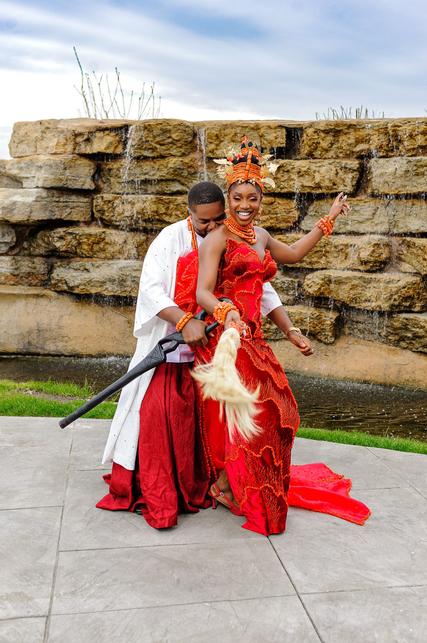 Nigerian bride and groom in traditional red and white attire pose playfully near stone waterfall.