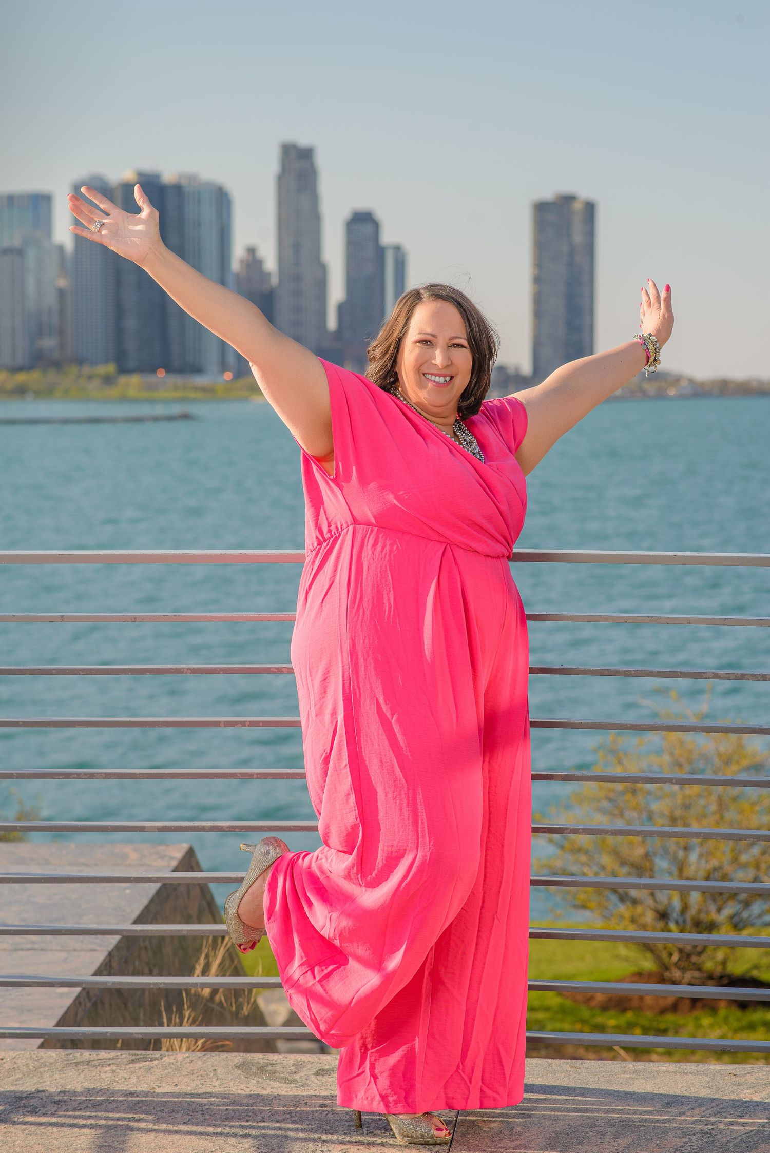 Someone in a flowing pink maxi dress poses joyfully with arms raised against Chicago skyline and Lake Michigan backdrop.