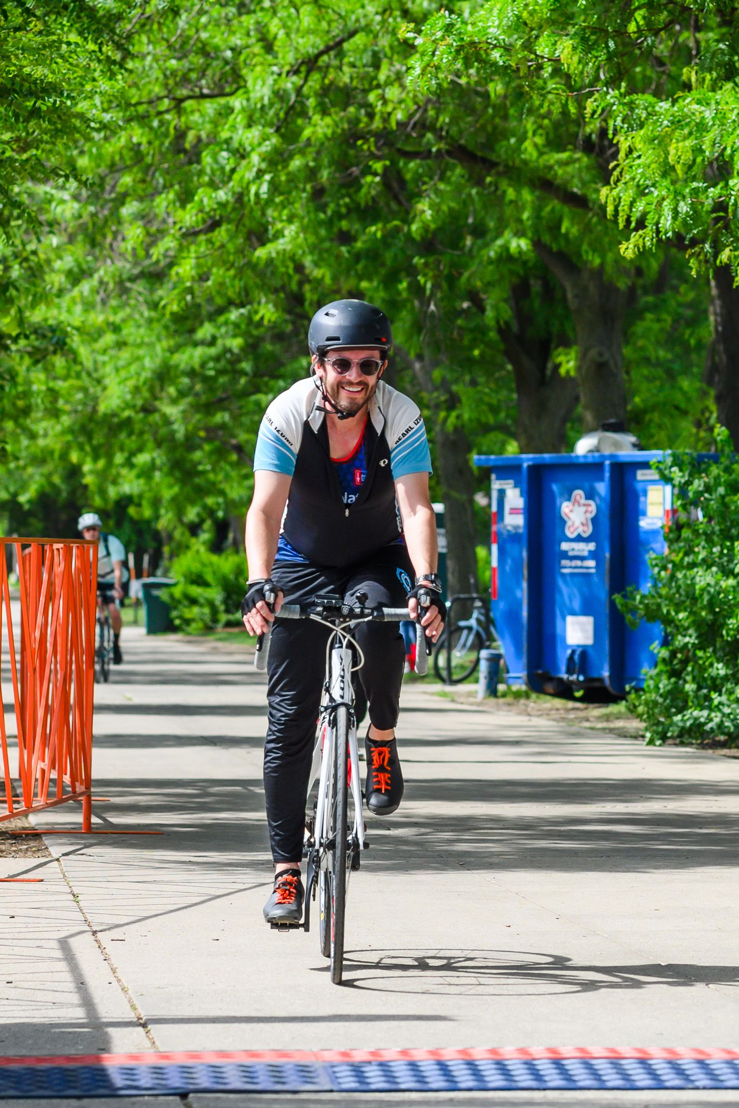 Smiling cyclist wearing helmet and sunglasses rides bicycle on tree-lined path during outdoor event.
