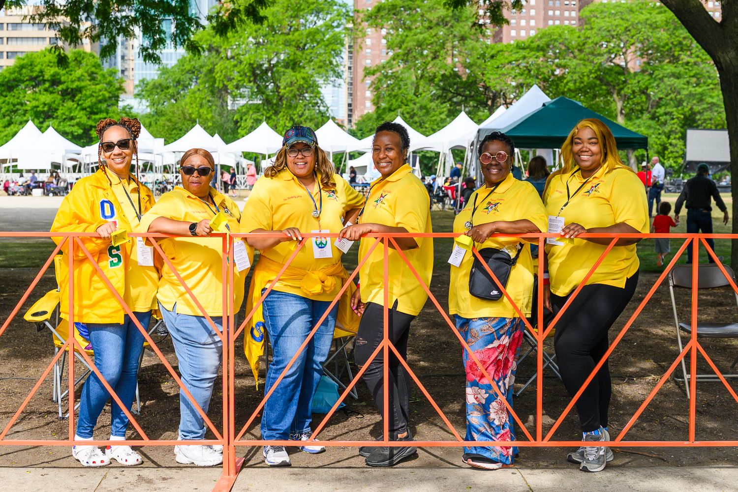 Six women in matching yellow shirts smile together at an outdoor festival with white tents and green trees.