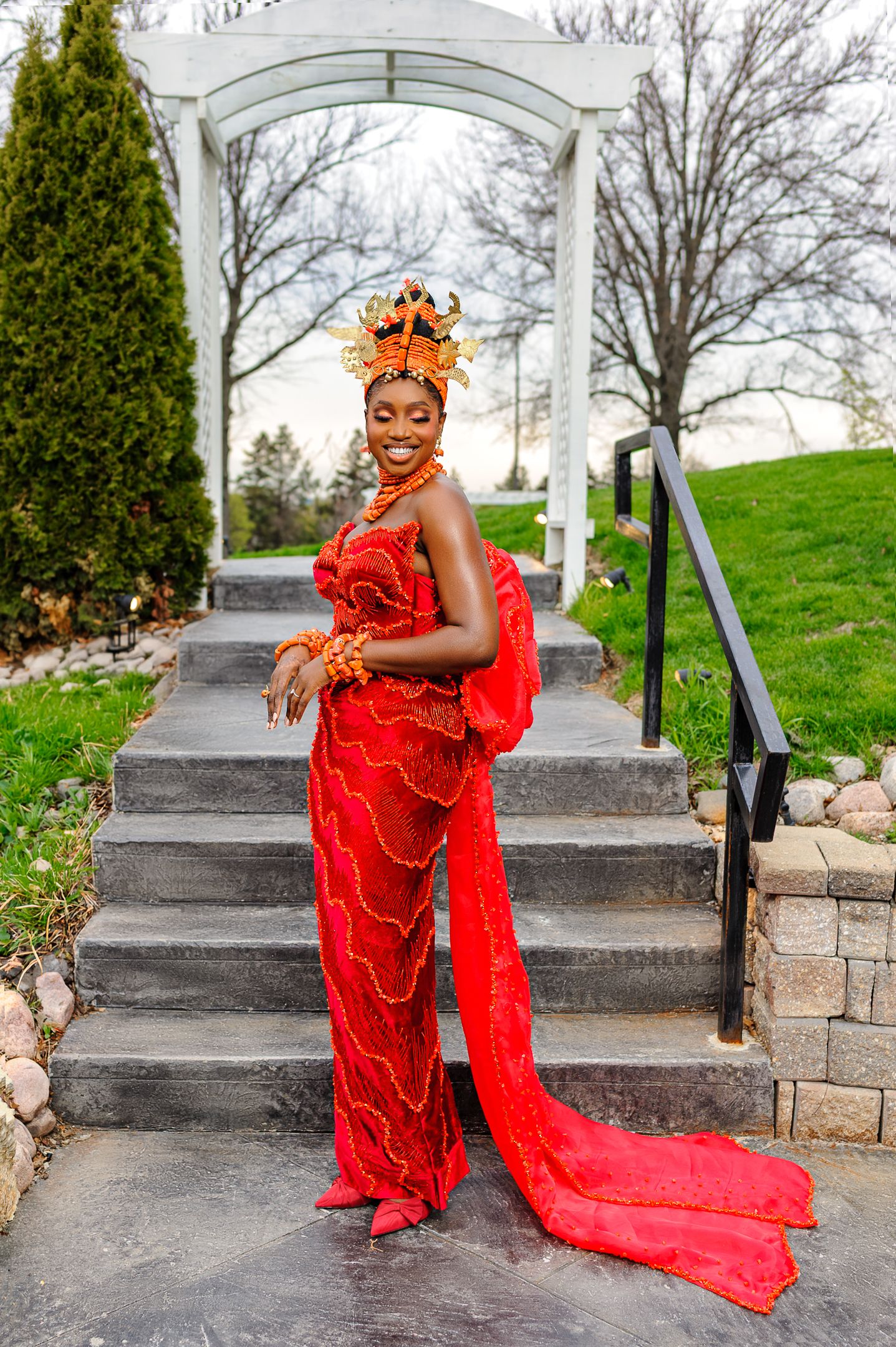 Smiling Nigerian bride in red traditional Urhobo wedding dress poses on stone steps at venue.