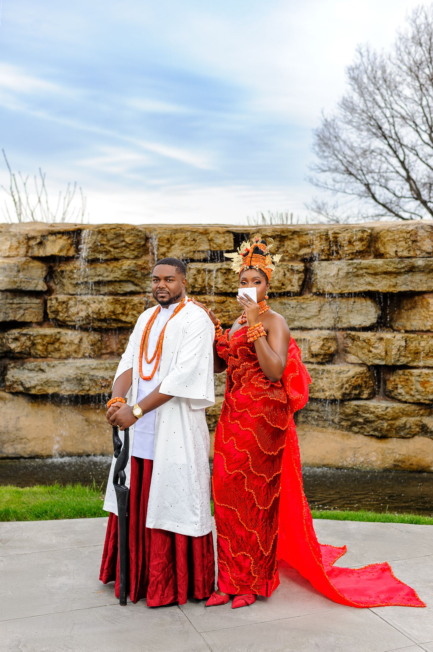 Couple in traditional Nigerian wedding attire, man in white and red, woman in red beaded gown with gold headdress.