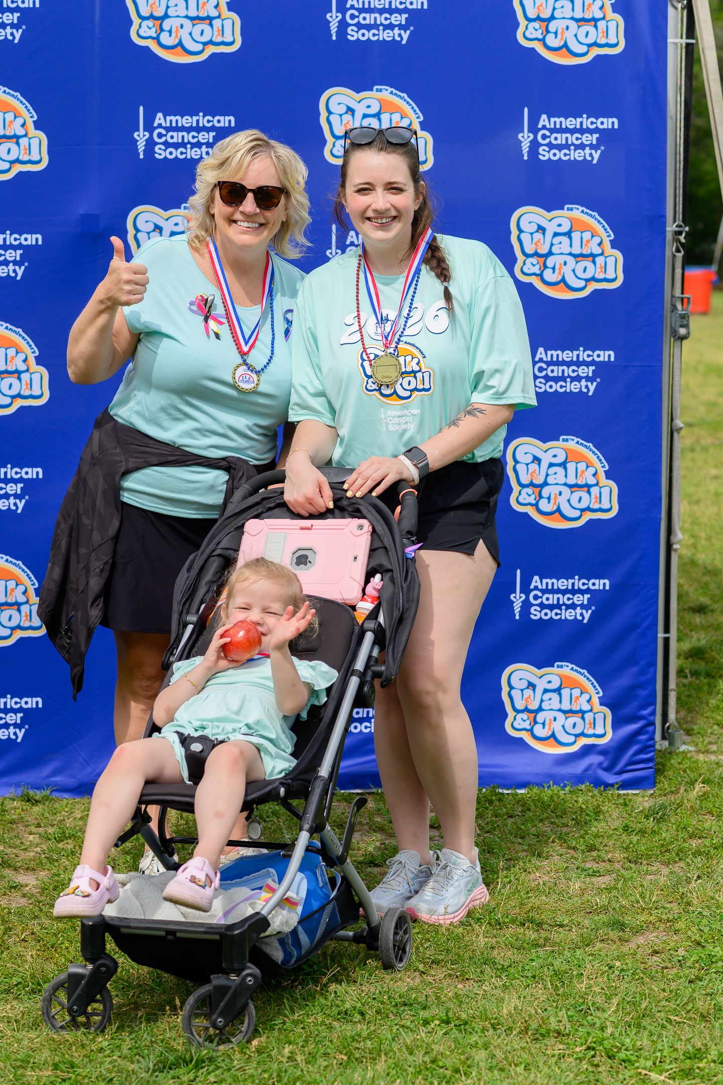 Two women wearing teal shirts and medals pose with a baby in a stroller at an American Cancer Society Walk & Roll event.