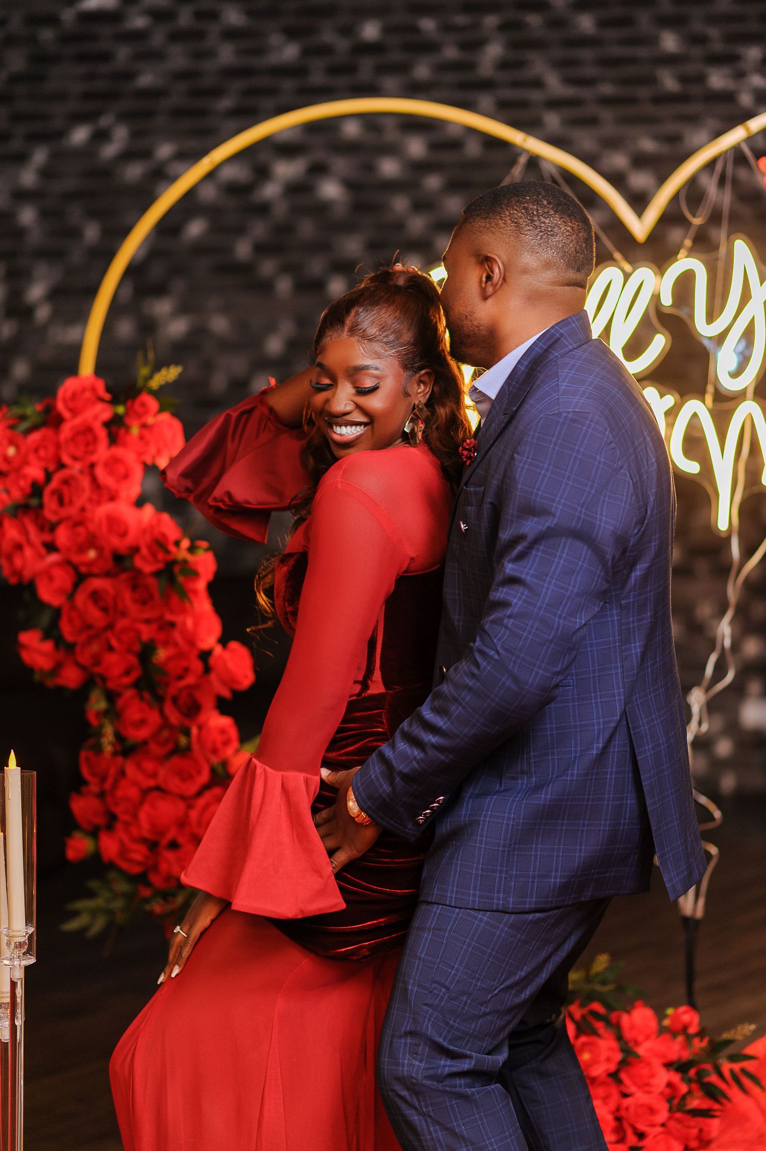 A joyful couple in red dress and blue suit embrace before a heart-shaped gold arch with red roses.