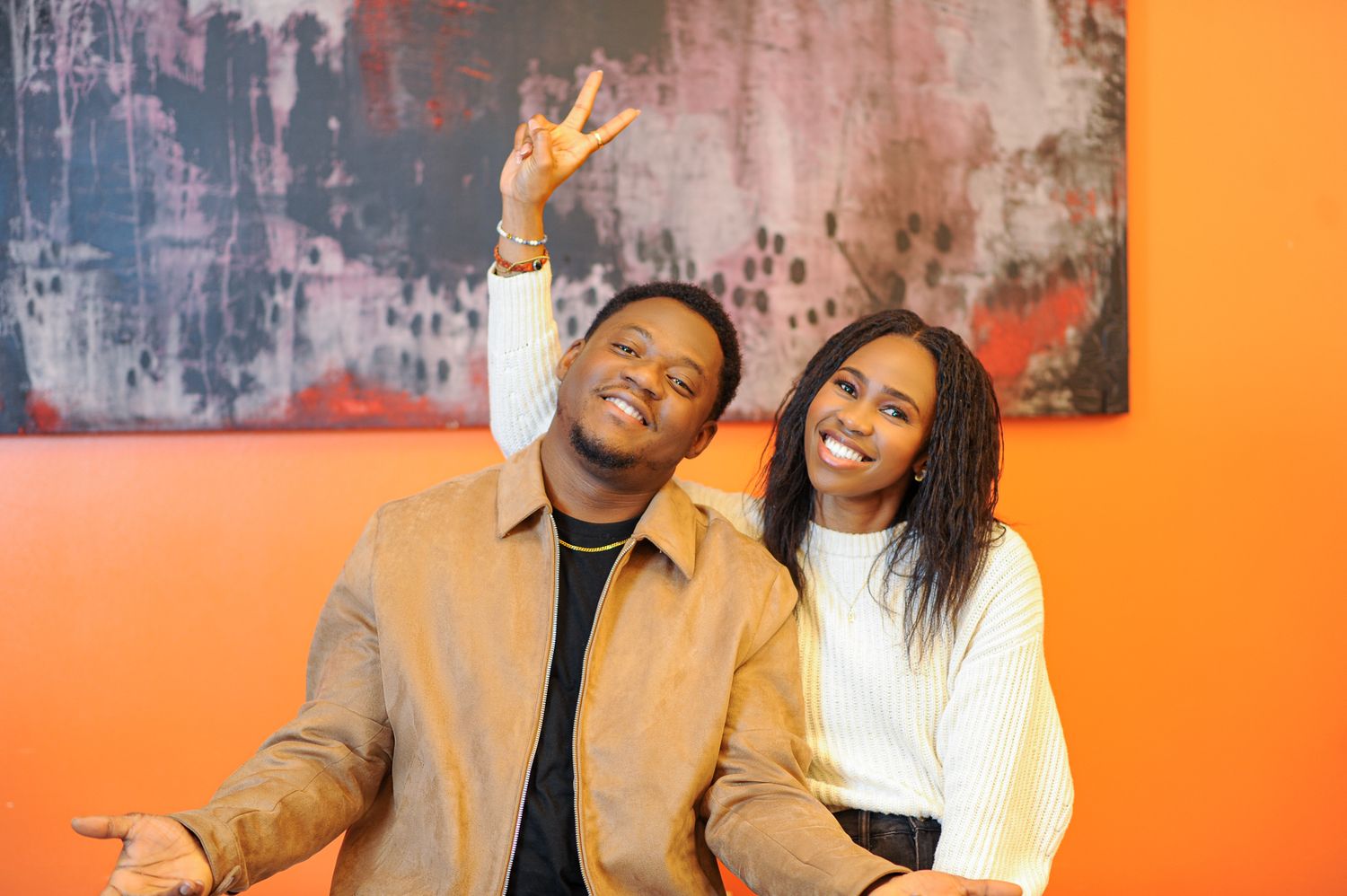 Two smiling friends pose together in front of an orange wall, one making a peace sign gesture.