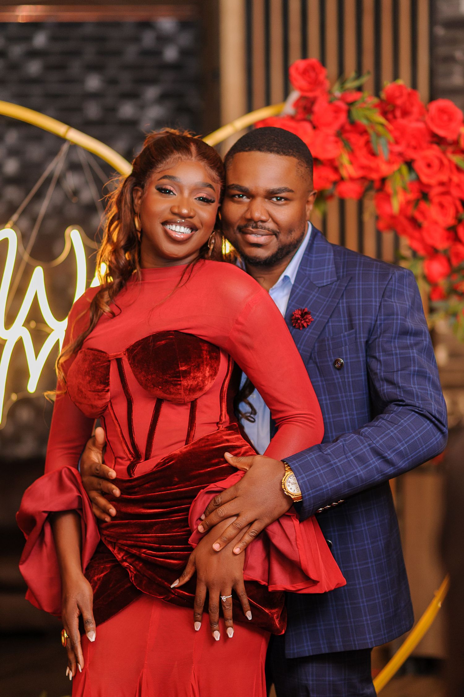 Couple posing elegantly, woman in red velvet gown and man in blue plaid suit, with red roses backdrop.