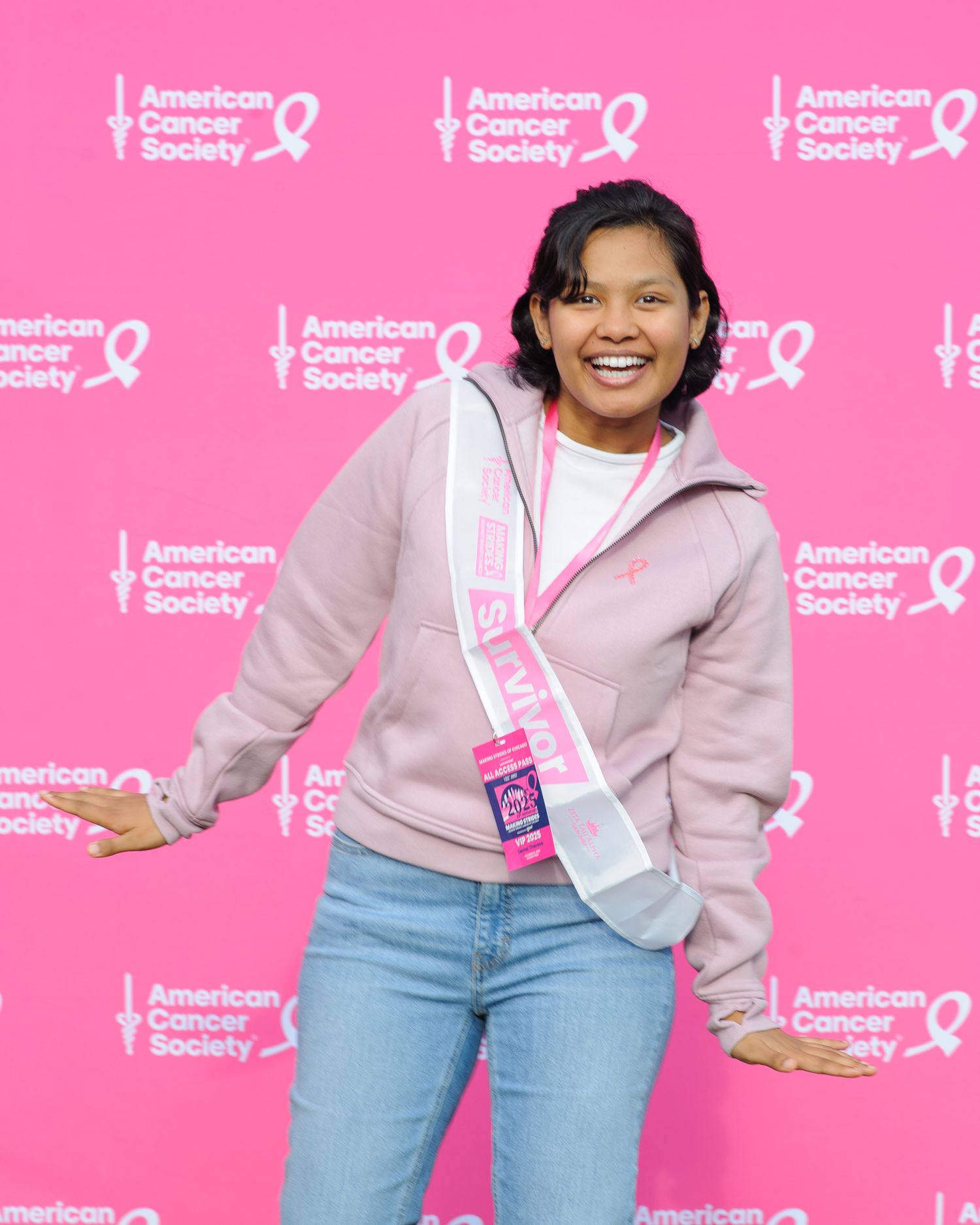 A person poses in casual attire against a pink backdrop with American Cancer Society logos and ribbons.