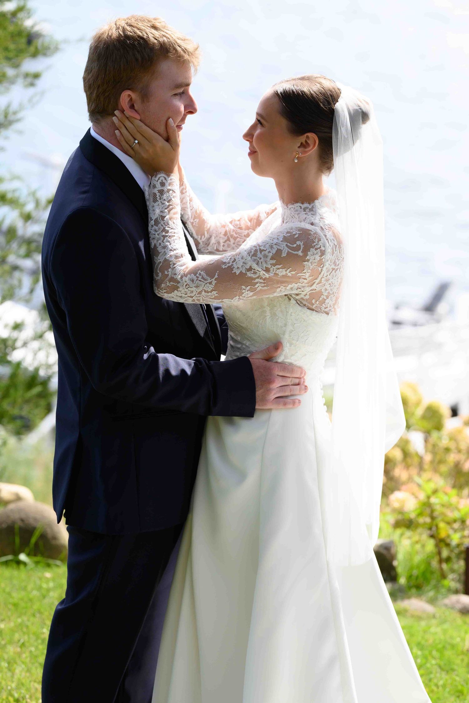 Bride in lace wedding gown and veil tenderly holds groom's face as they share a romantic moment outdoors.