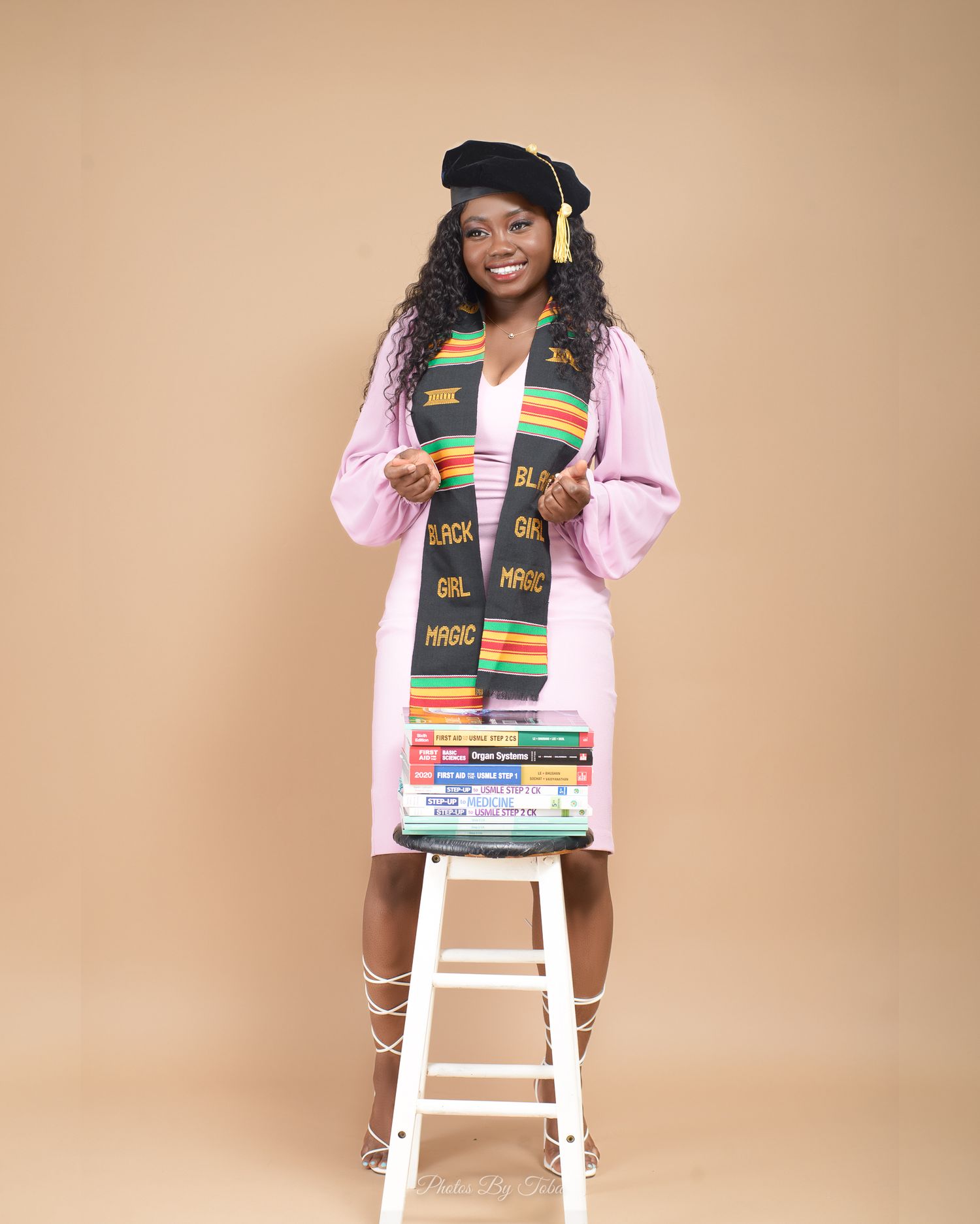 Graduate in academic regalia with a colorful kente stole poses on white stool against neutral backdrop.