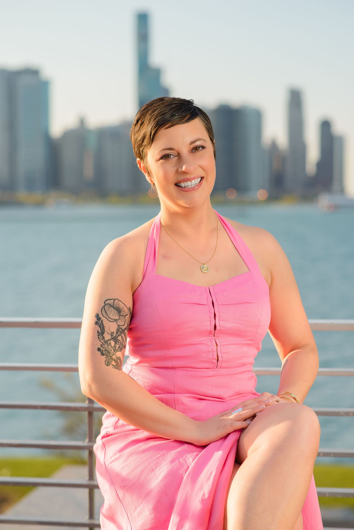 Person in pink dress sitting by waterfront with city skyline in background on sunny day.