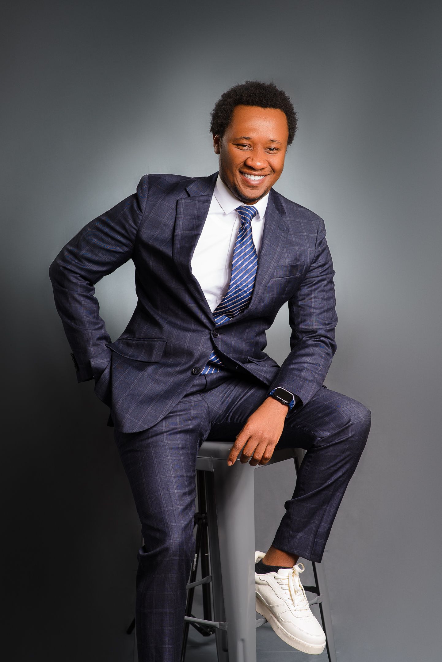Professional wearing a navy pinstripe suit and striped tie poses on a stool against grey backdrop in white sneakers.