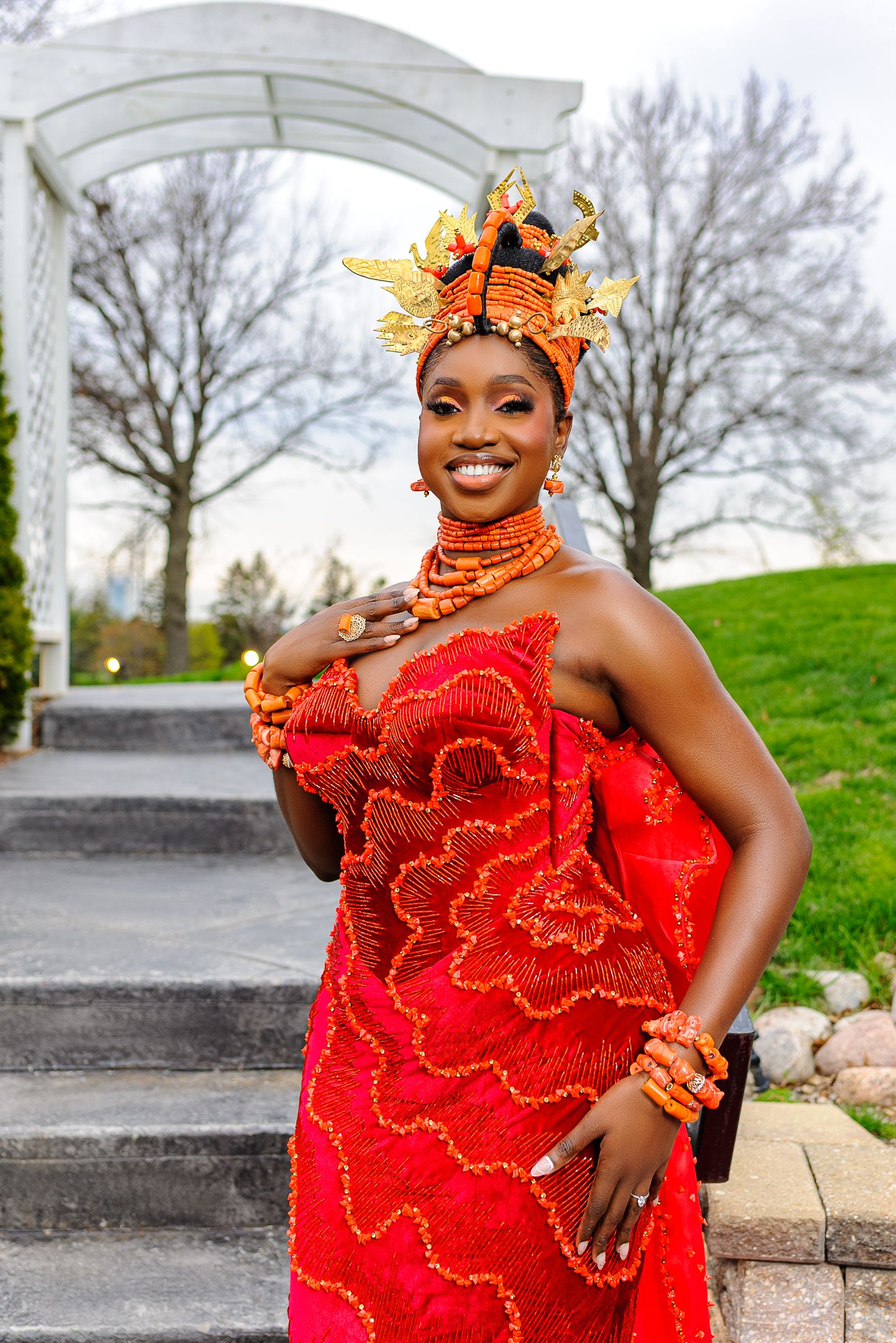 Woman in vibrant red African traditional attire with ornate orange headdress and beaded jewelry smiling outdoors.
