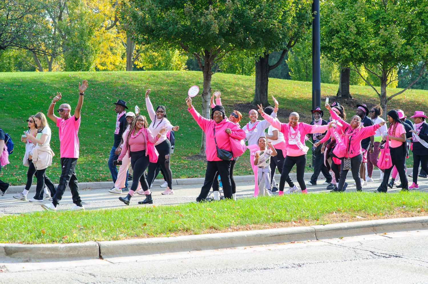 A large group of people in pink and black outfits exercise together outdoors in a park setting.