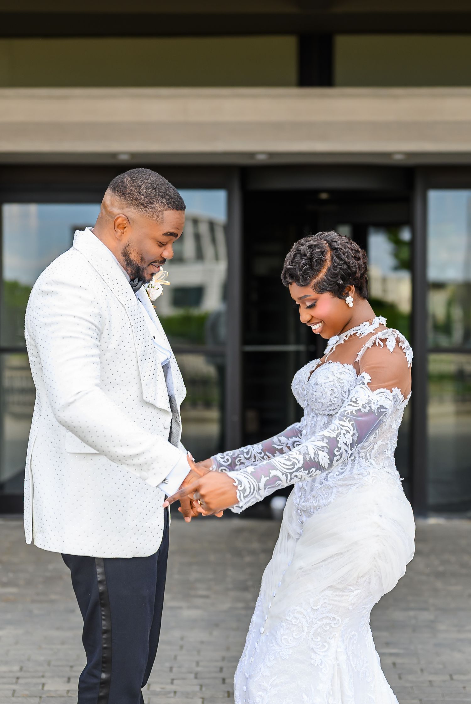 A bride and groom hold hands outdoors, she in lace wedding gown, he in white suit jacket, sharing a joyful moment.