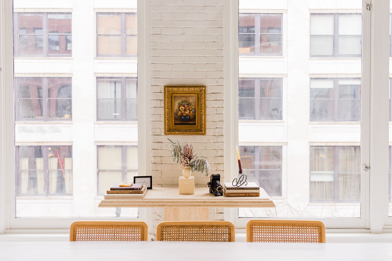 Minimalist white interior features exposed brick walls, high windows, and wooden dining chairs lined along a wall.