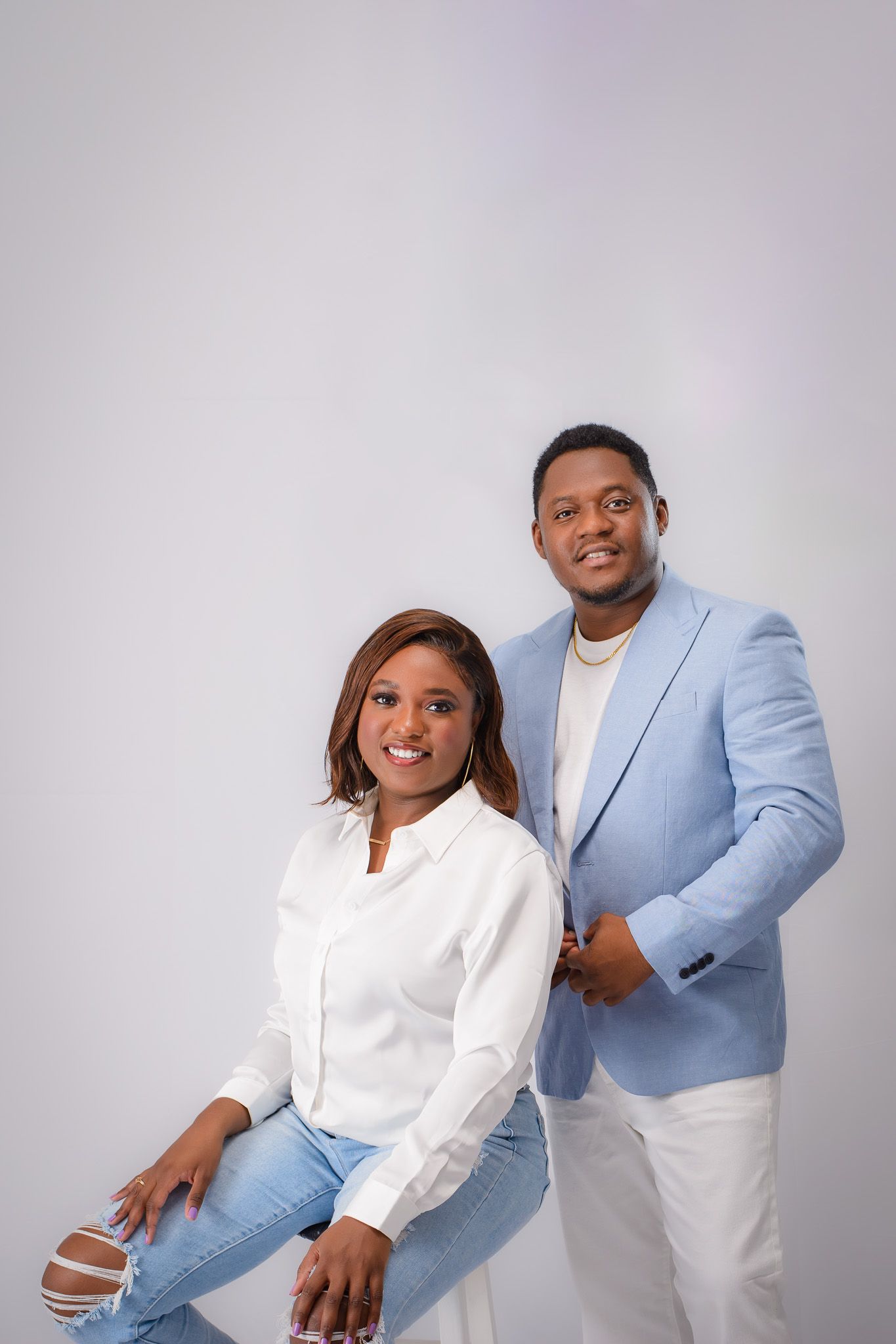 Studio portrait of couple in light blue suit and white outfit against grey background.