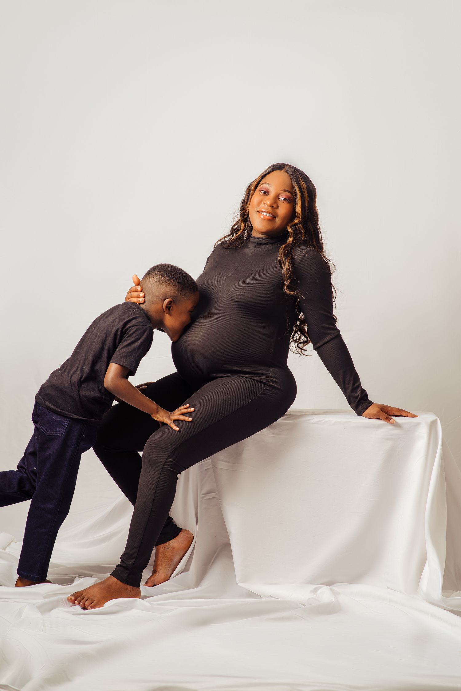 Sweet maternity session with child kissing pregnant belly on white backdrop.