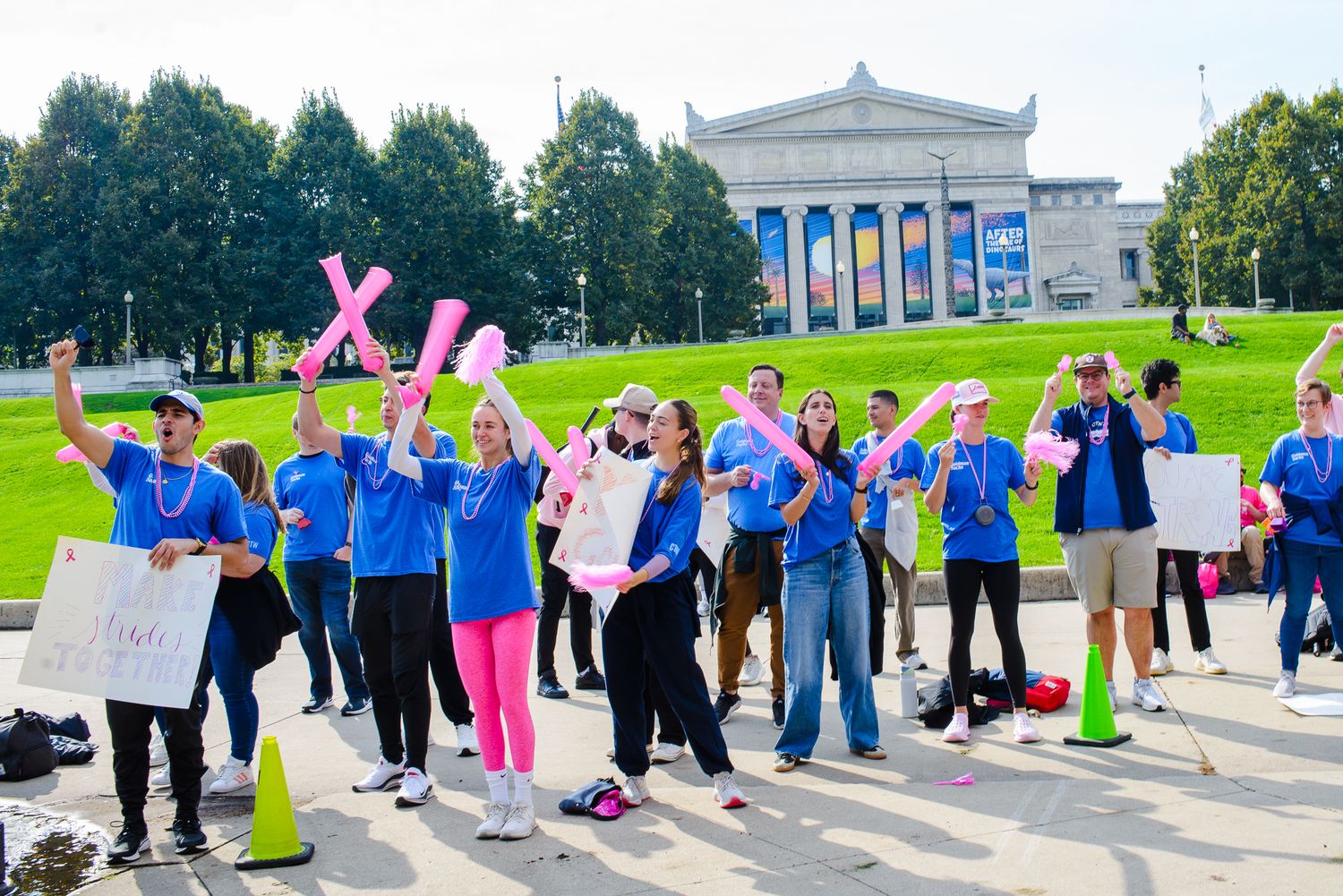A group of volunteers in blue jackets gather with signs and pink X symbols outside a neoclassical museum building.