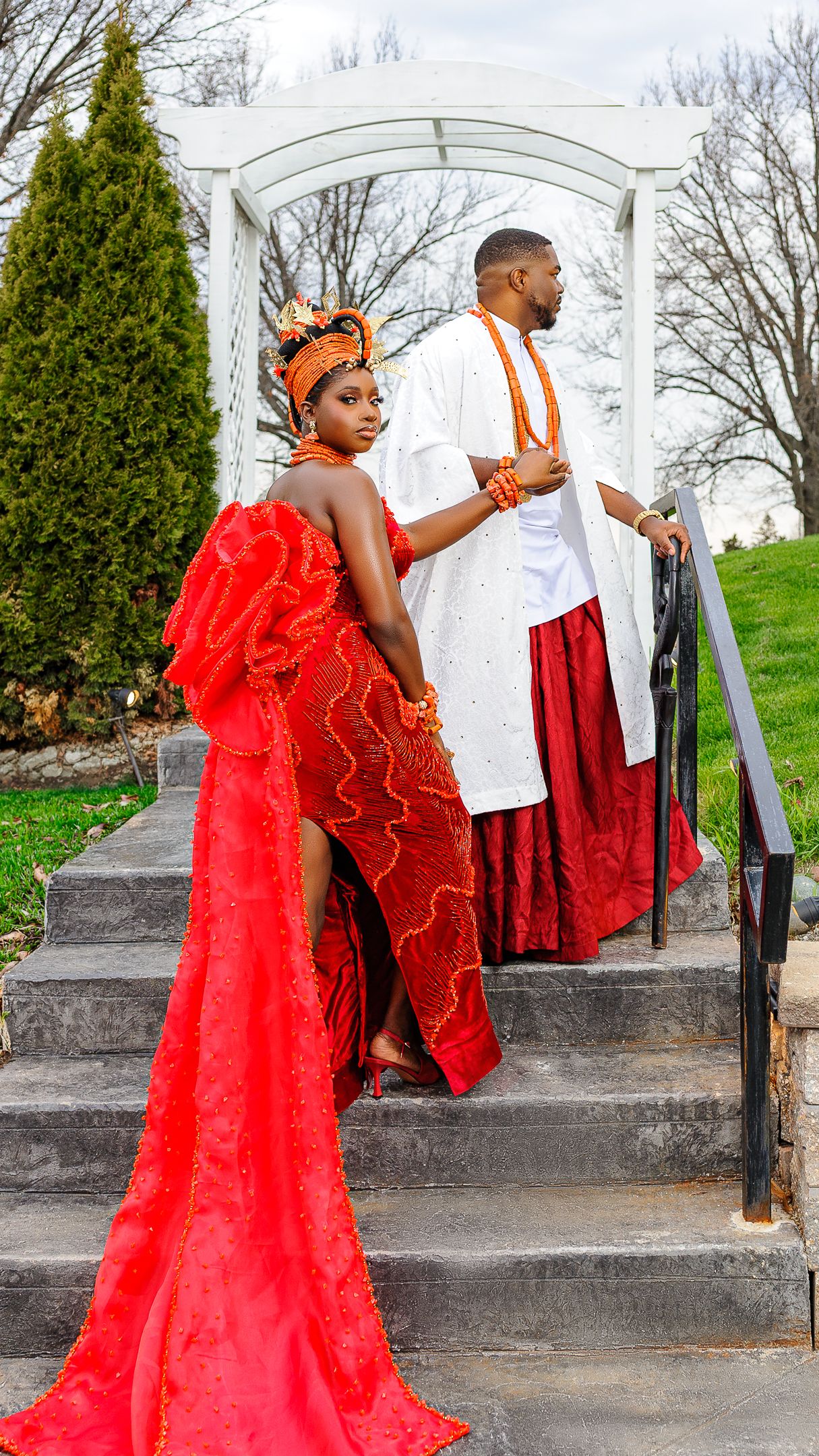 Couple in vibrant Nigerian traditional wedding attire, woman in red gown with train, man in white and red agbada, on steps.
