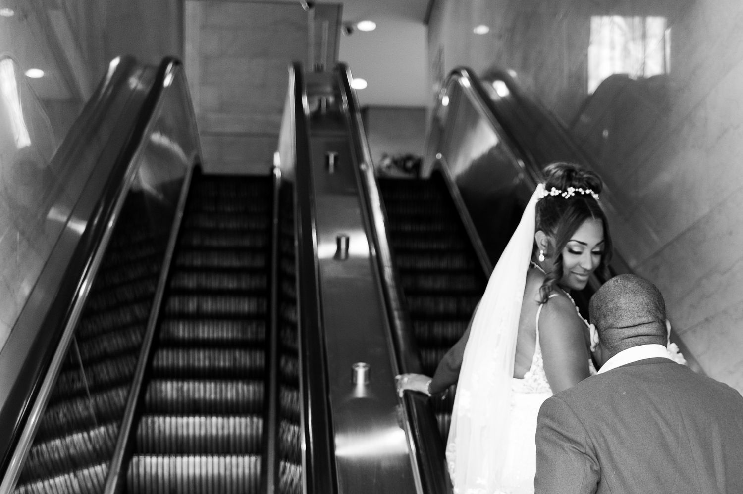 Black and white photo of a bride and groom riding an escalator on their wedding day.