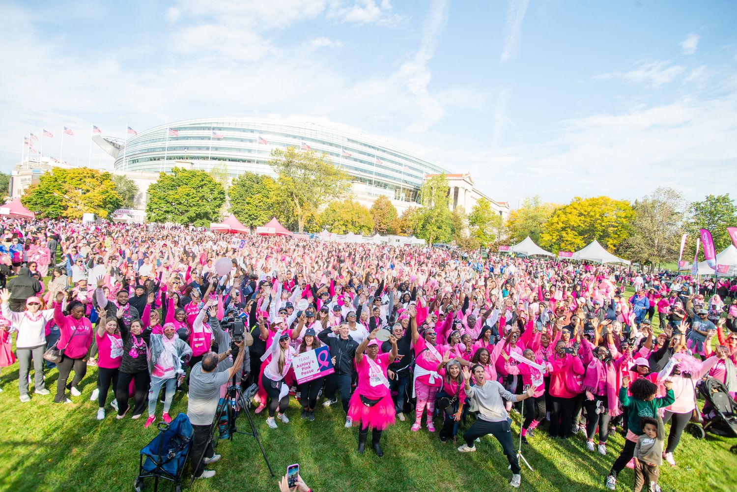 A large crowd of participants wearing pink shirts gather outdoors for a breast cancer awareness event or charity walk.