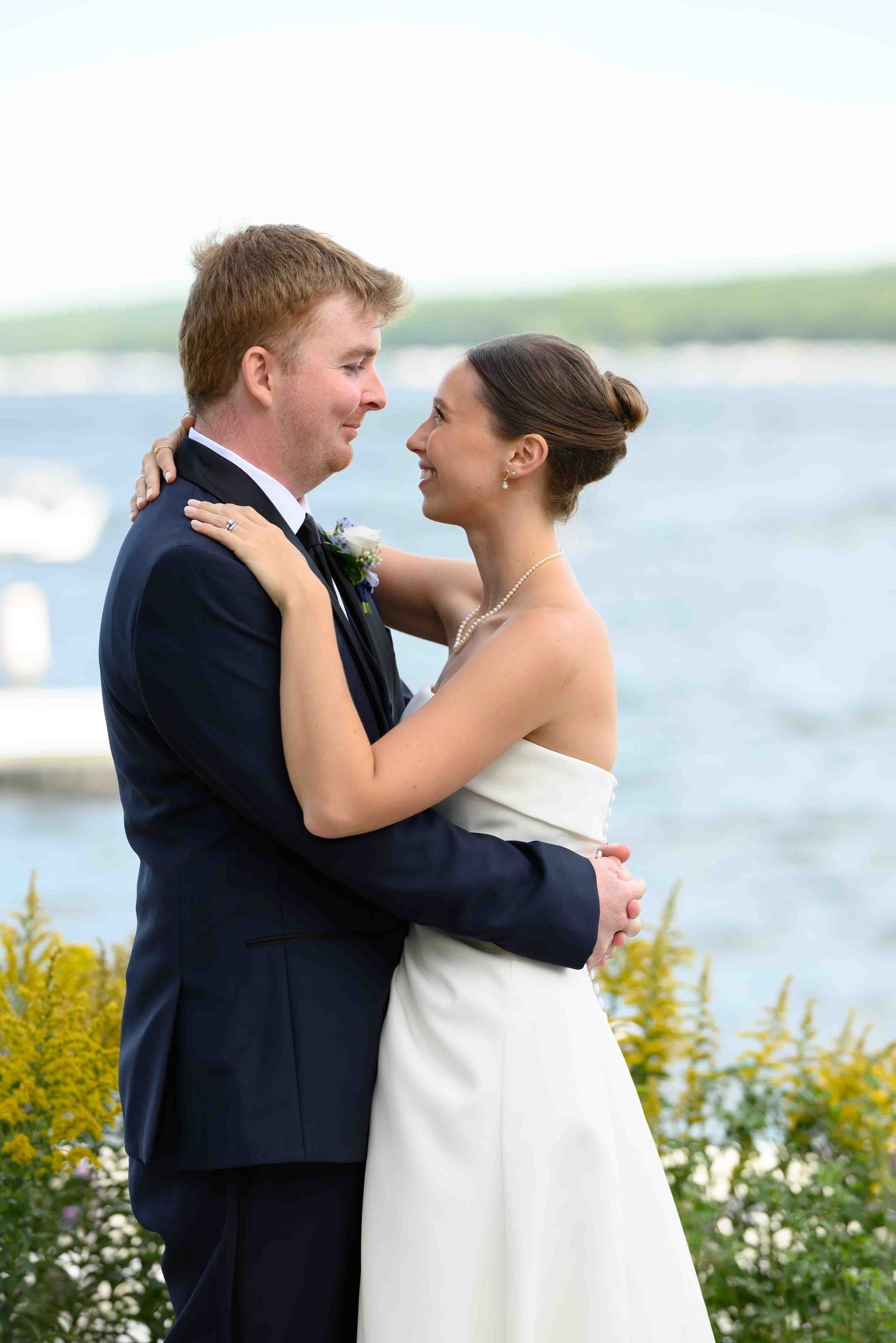 Bride and groom embracing outdoors near waterfront, smiling at each other, wearing wedding attire with flowers in background.