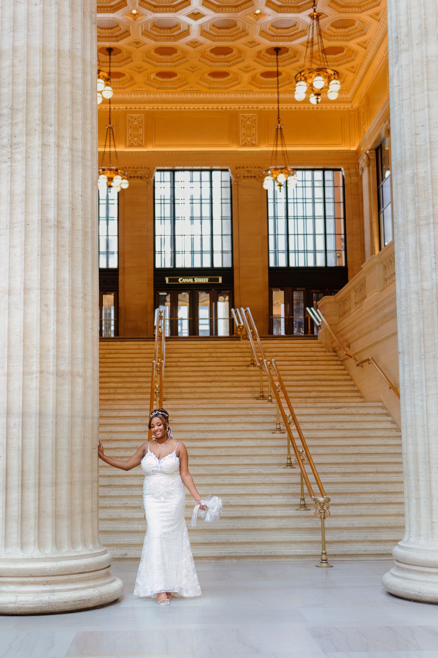 Bride in white lace gown poses on grand staircase of elegant Union Station with tall columns and ornate ceiling.