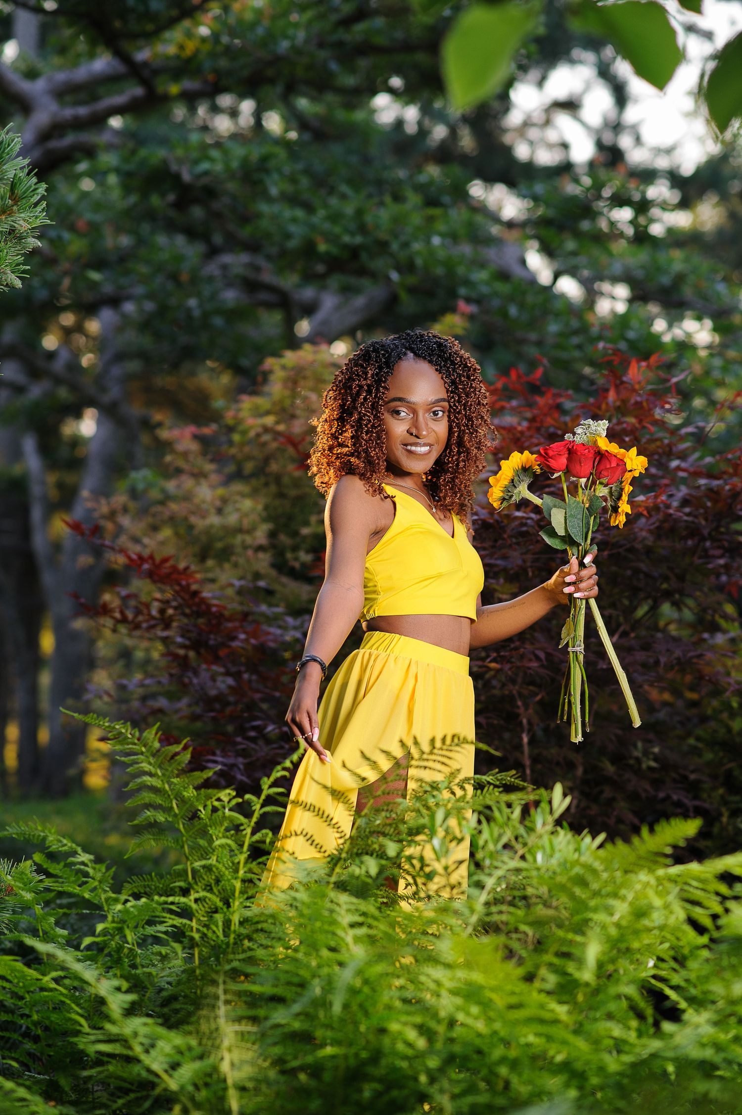Person in bright yellow dress holding colorful sunflowers while standing among lush green ferns in a garden setting.