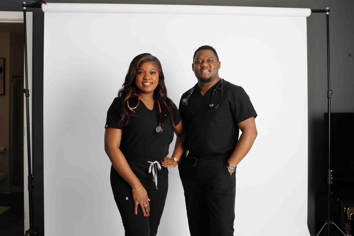 Two healthcare professionals in black scrubs with stethoscopes pose smiling in front of a white backdrop.