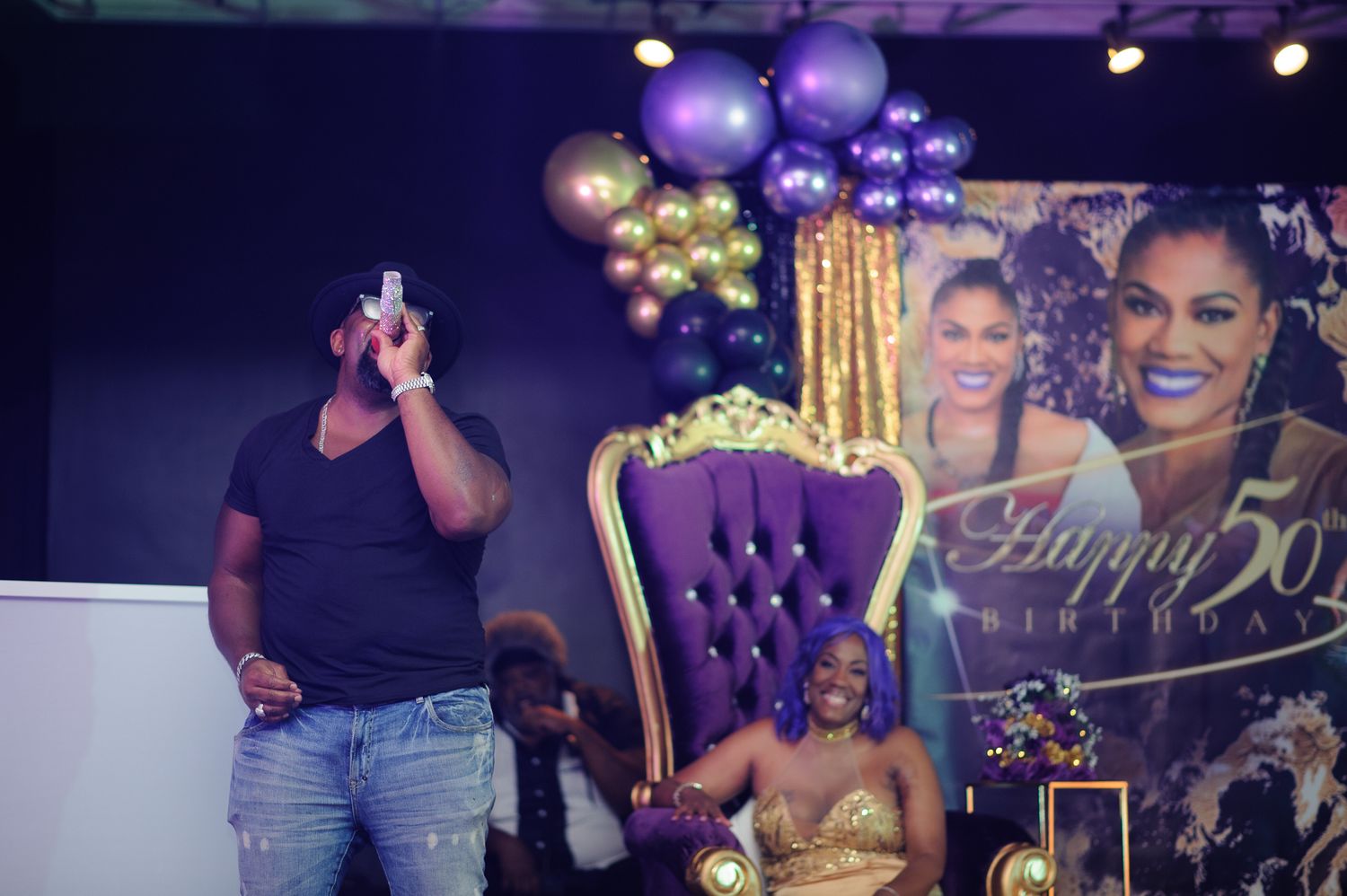 A woman sits on a purple throne chair at a Happy 50th Birthday party with gold and purple balloon decorations.