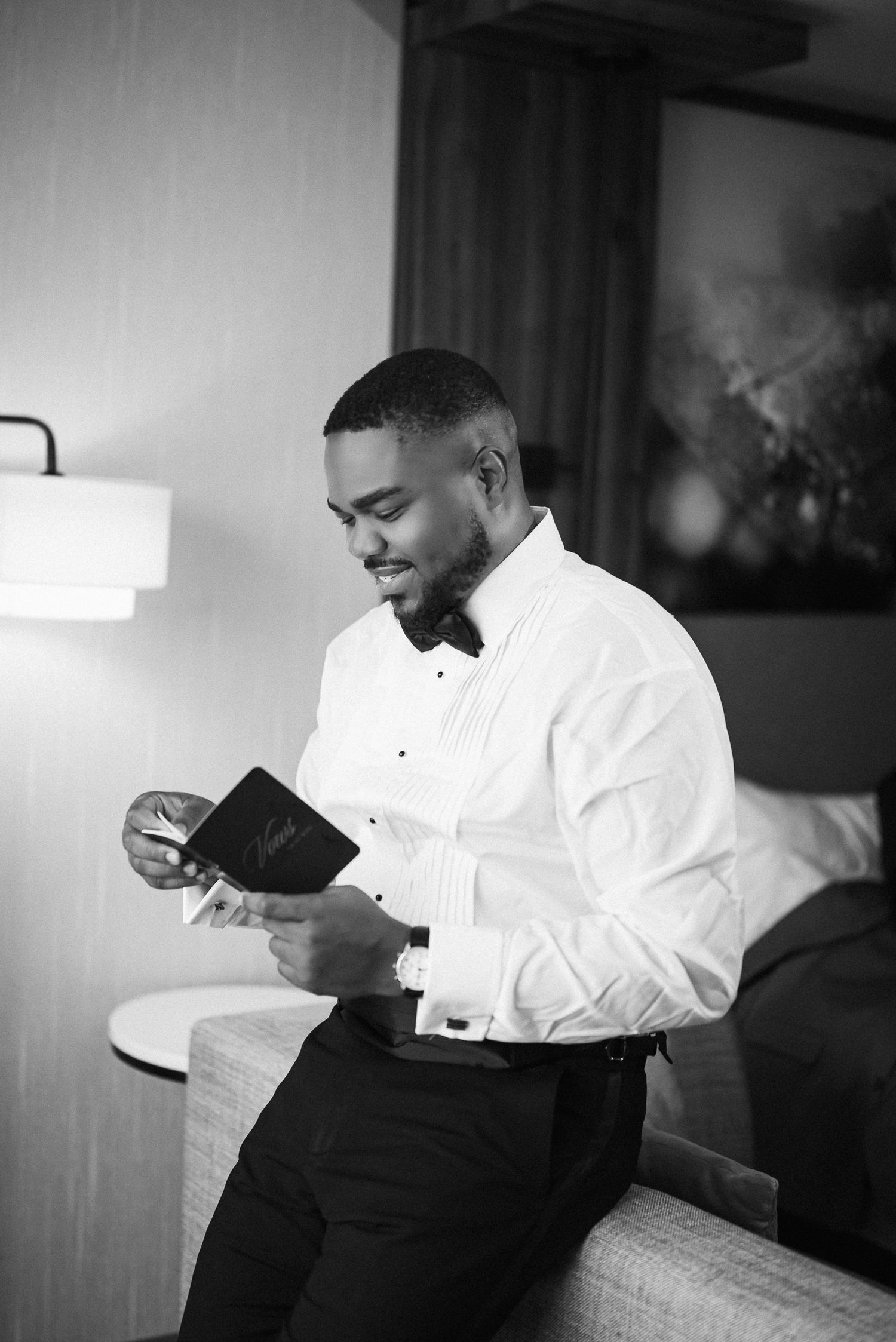 Black and white photo of a well-dressed groom in a tuxedo shirt and bow tie reading a card while seated.