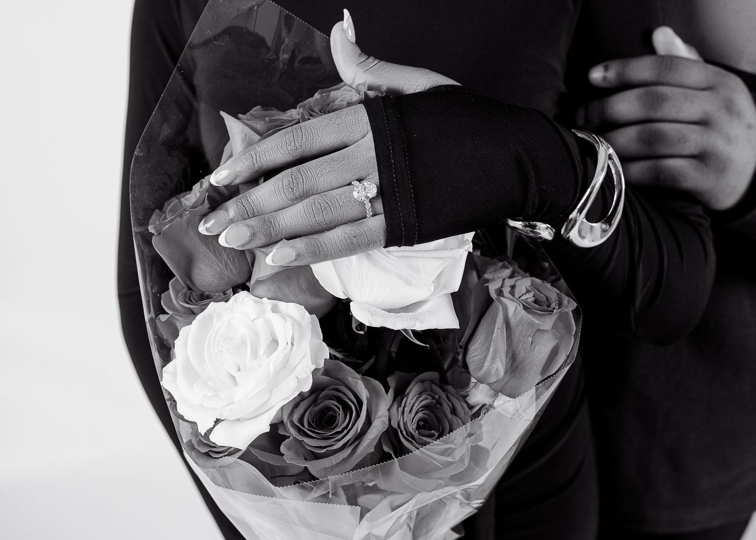 Black and white photo of couple embracing, showing engagement ring and bracelet over a bouquet of roses.