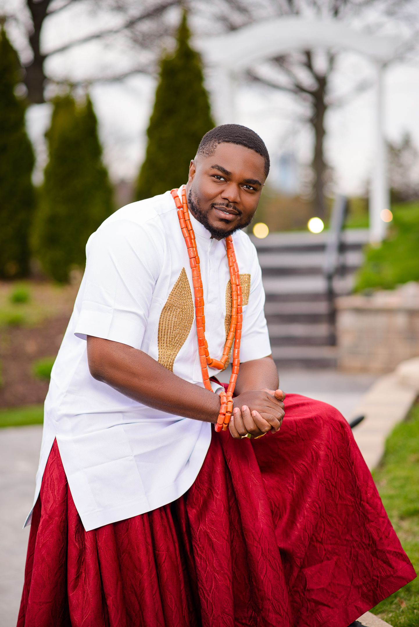 Nigerian groom in white traditional top and red wrapper with coral beads sits outdoors.