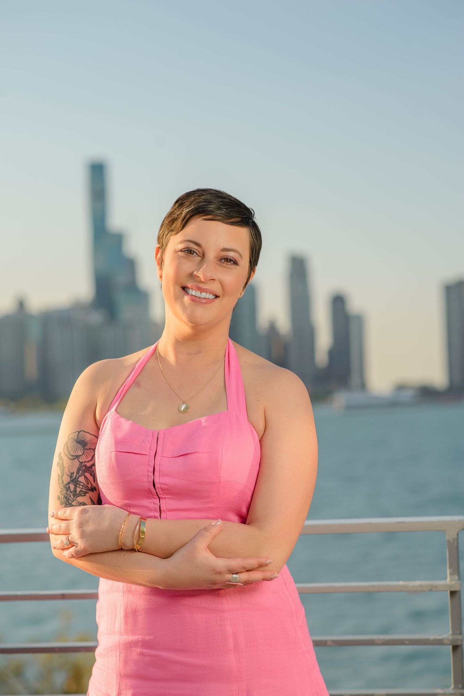 Person in pink tank top poses by waterfront railing with city skyline in background at sunset.