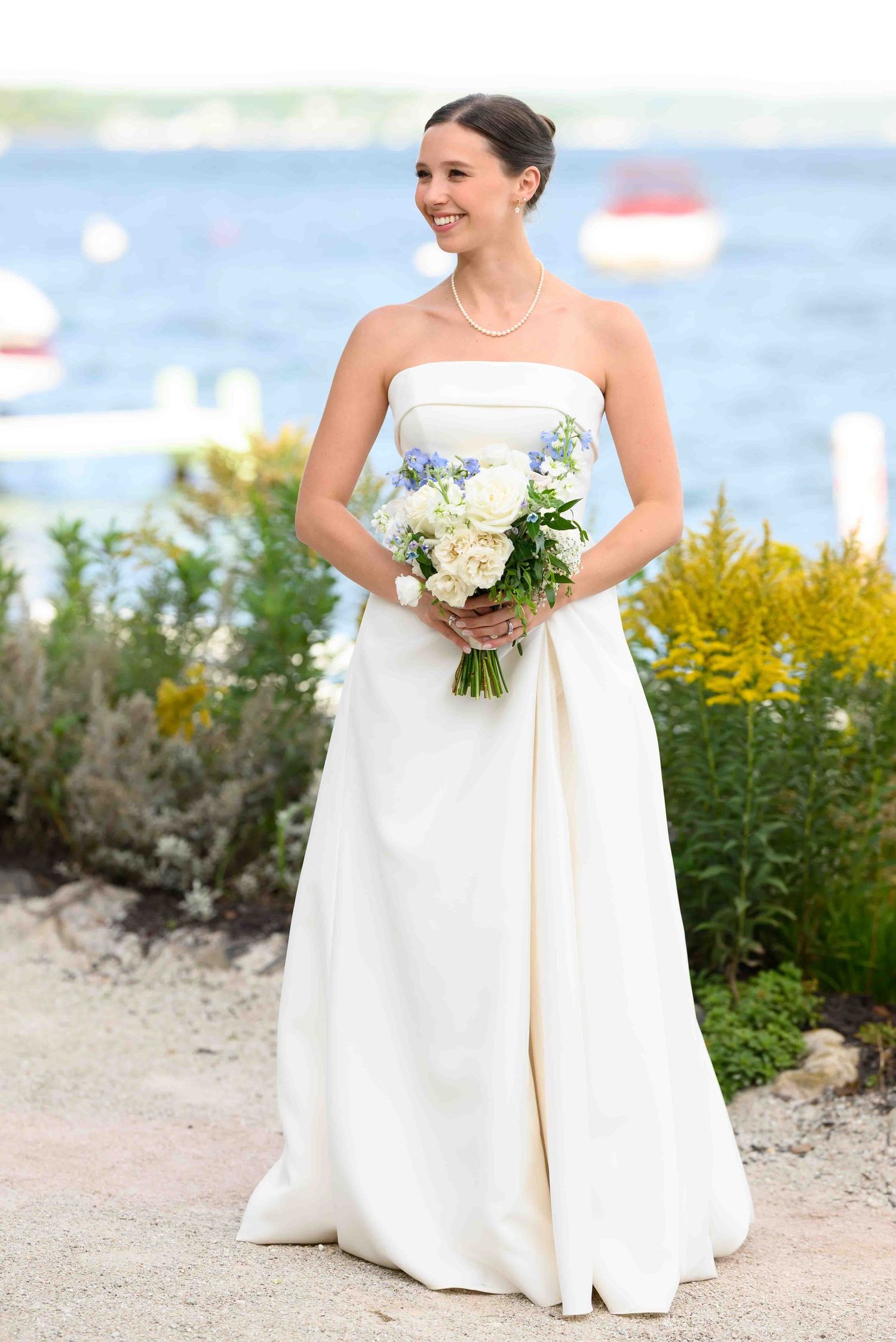 Smiling bride in strapless white gown holding blue and white bouquet, standing near waterfront with lush greenery.